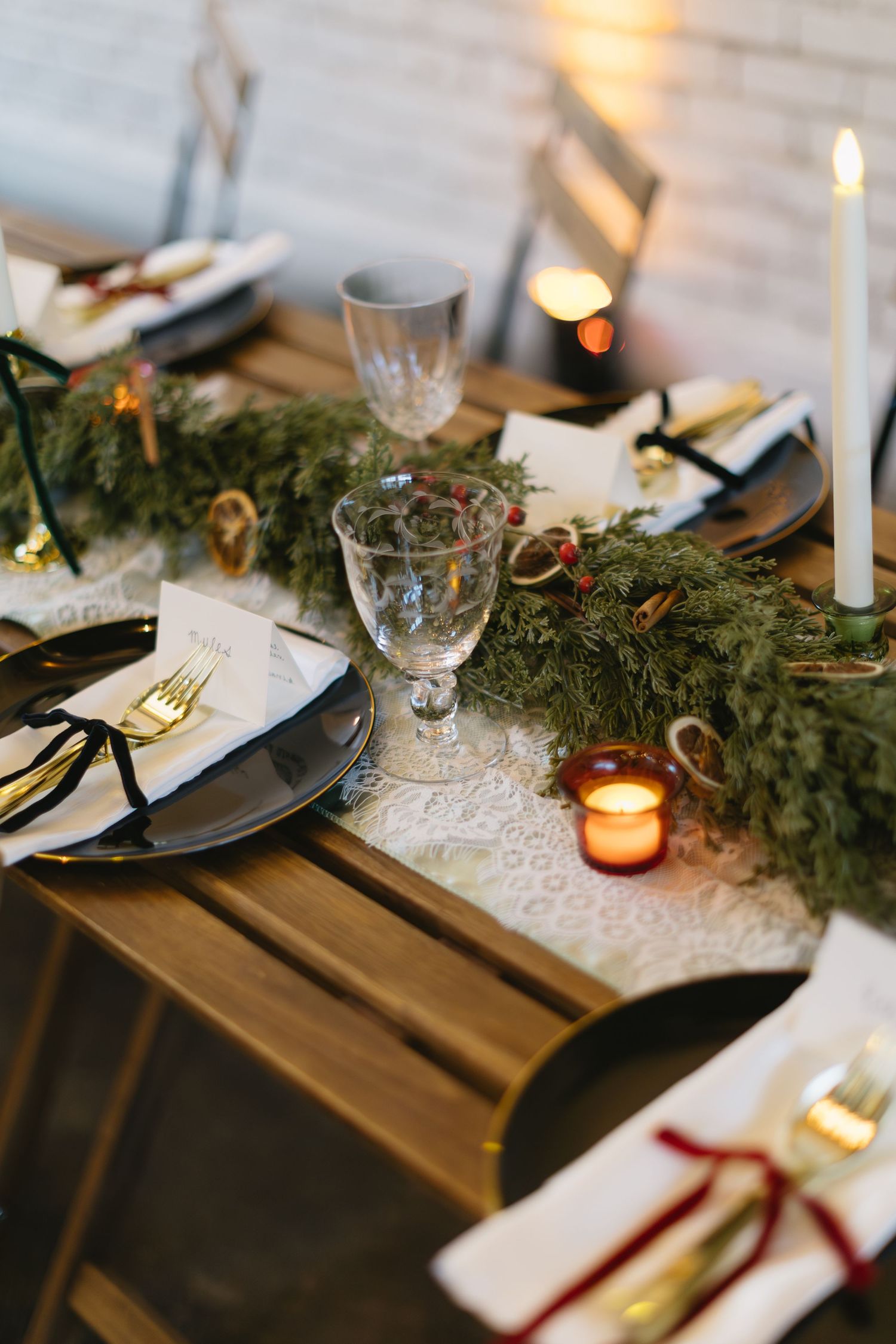 Elegant holiday table setting with evergreen garland, white candles, and gold cutlery on dark wooden table.
