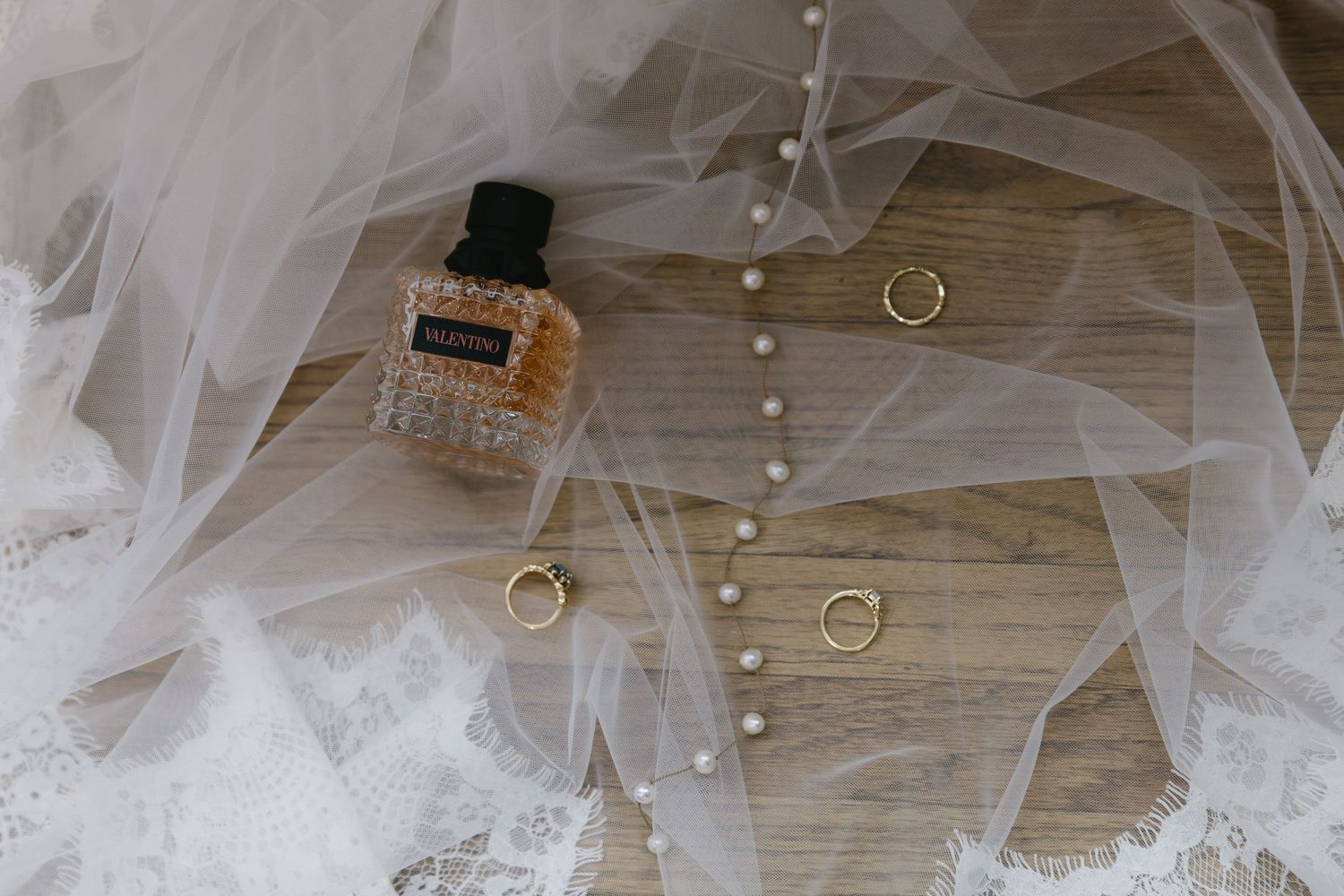 Wedding details including perfume bottle, delicate veil tulle, and gold rings arranged on wooden surface.