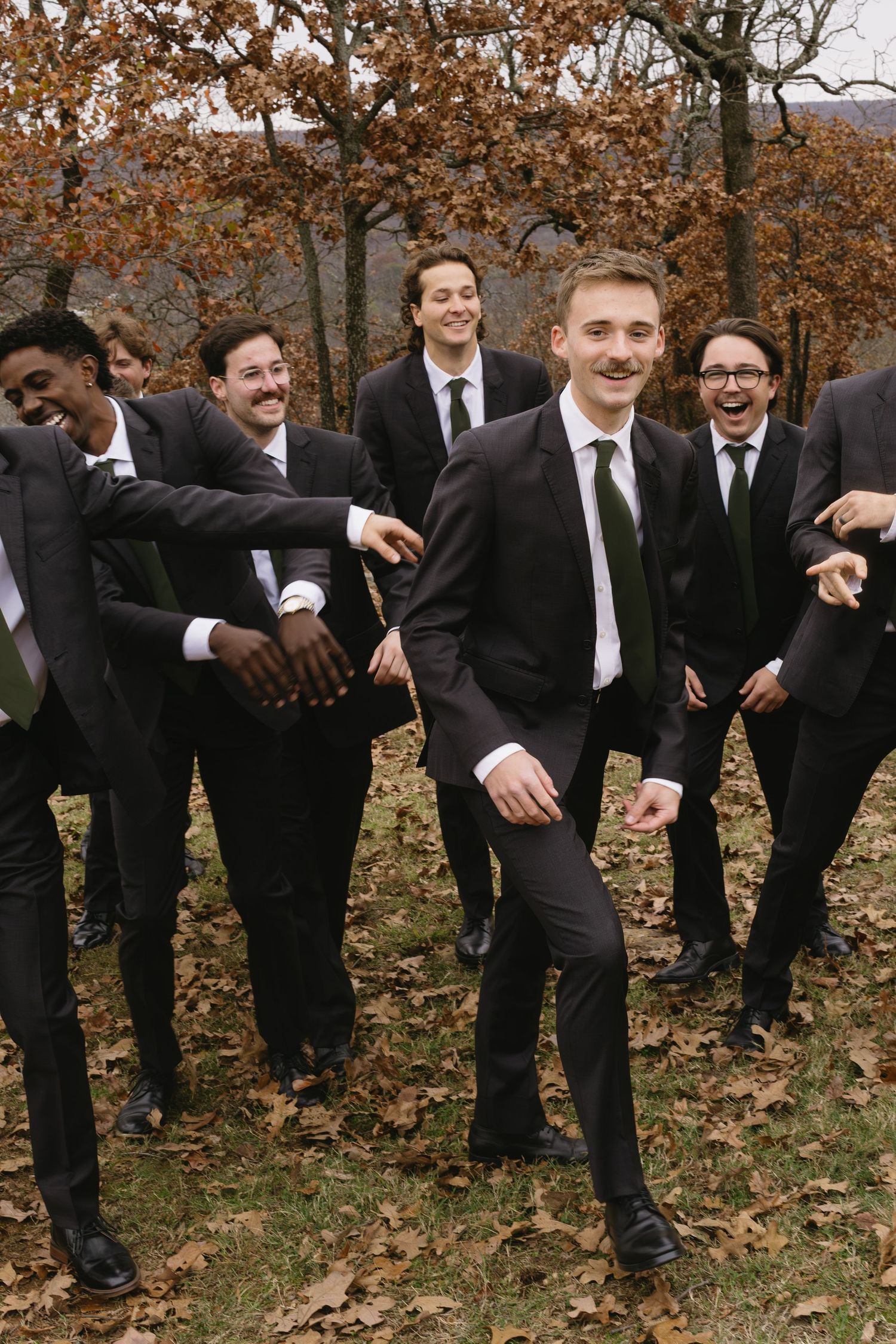 Groomsmen in black suits and ties strike playful poses in a line during autumn wedding photos.