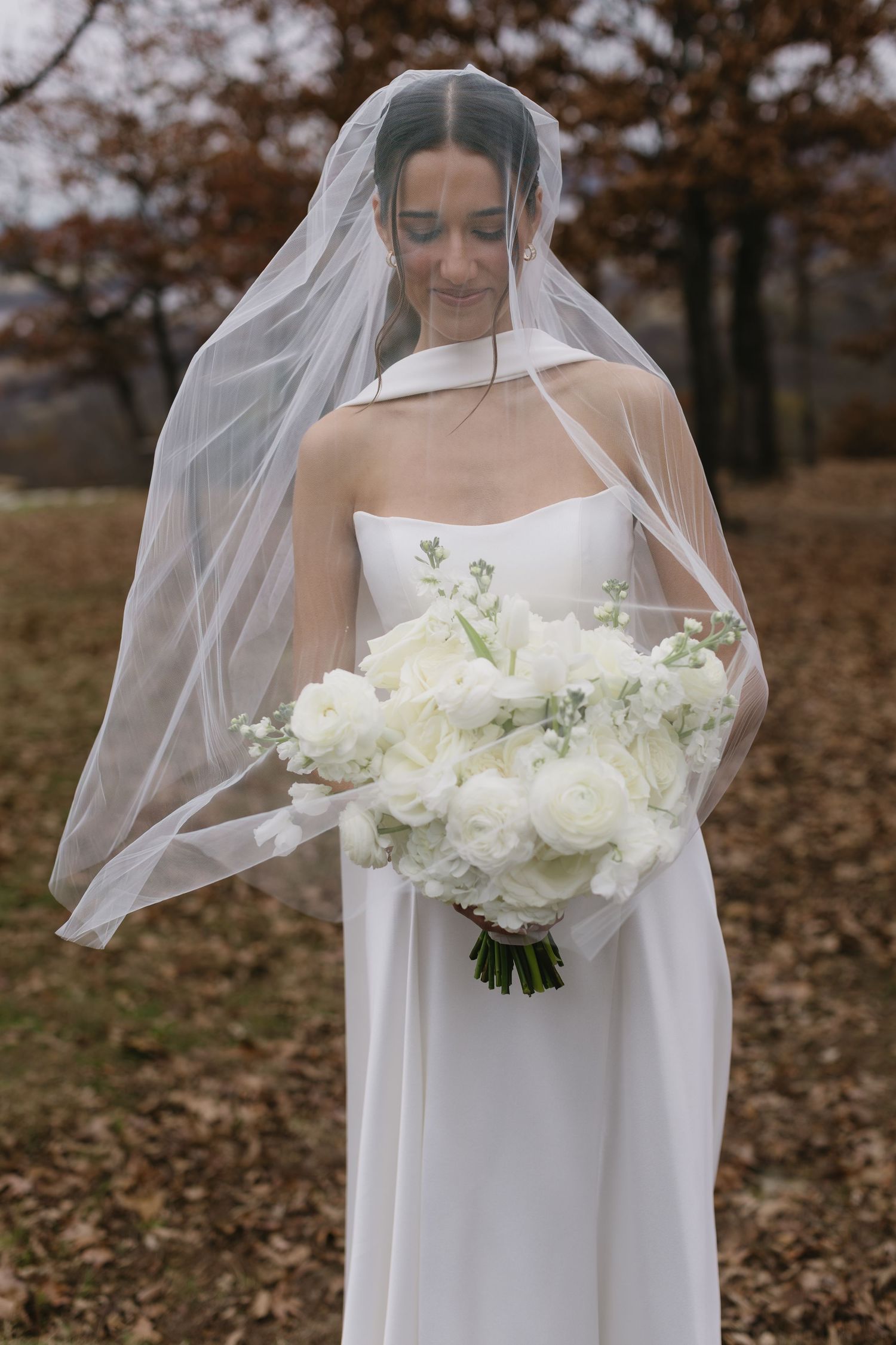 Bride in flowing white dress holds elegant white floral bouquet in autumn forest.