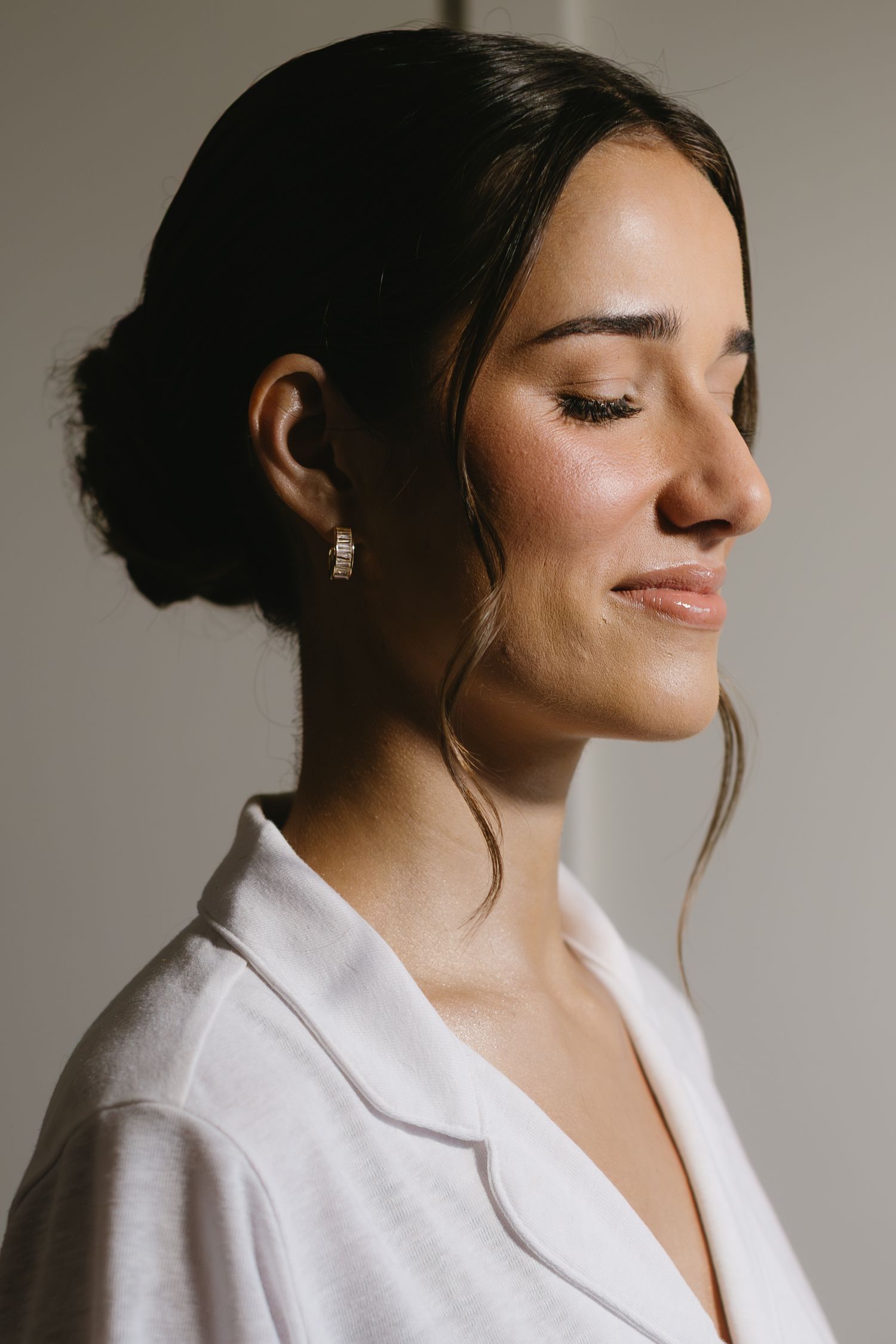 Side profile portrait of someone in white wearing delicate earrings against a neutral background.