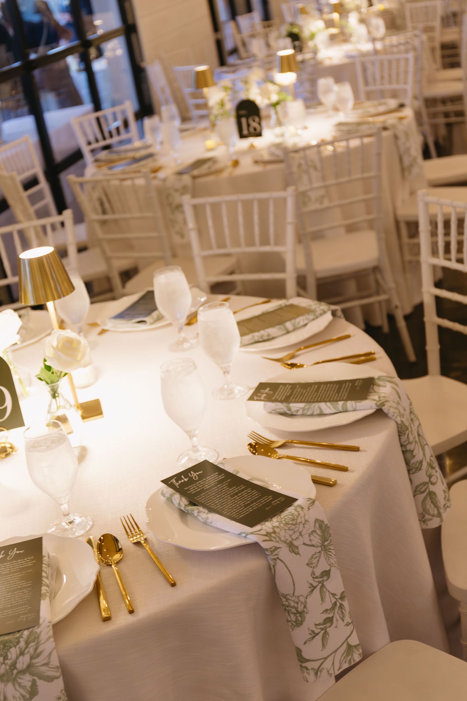 Elegant wedding reception table setting with white linens, gold cutlery, and white Chiavari chairs.