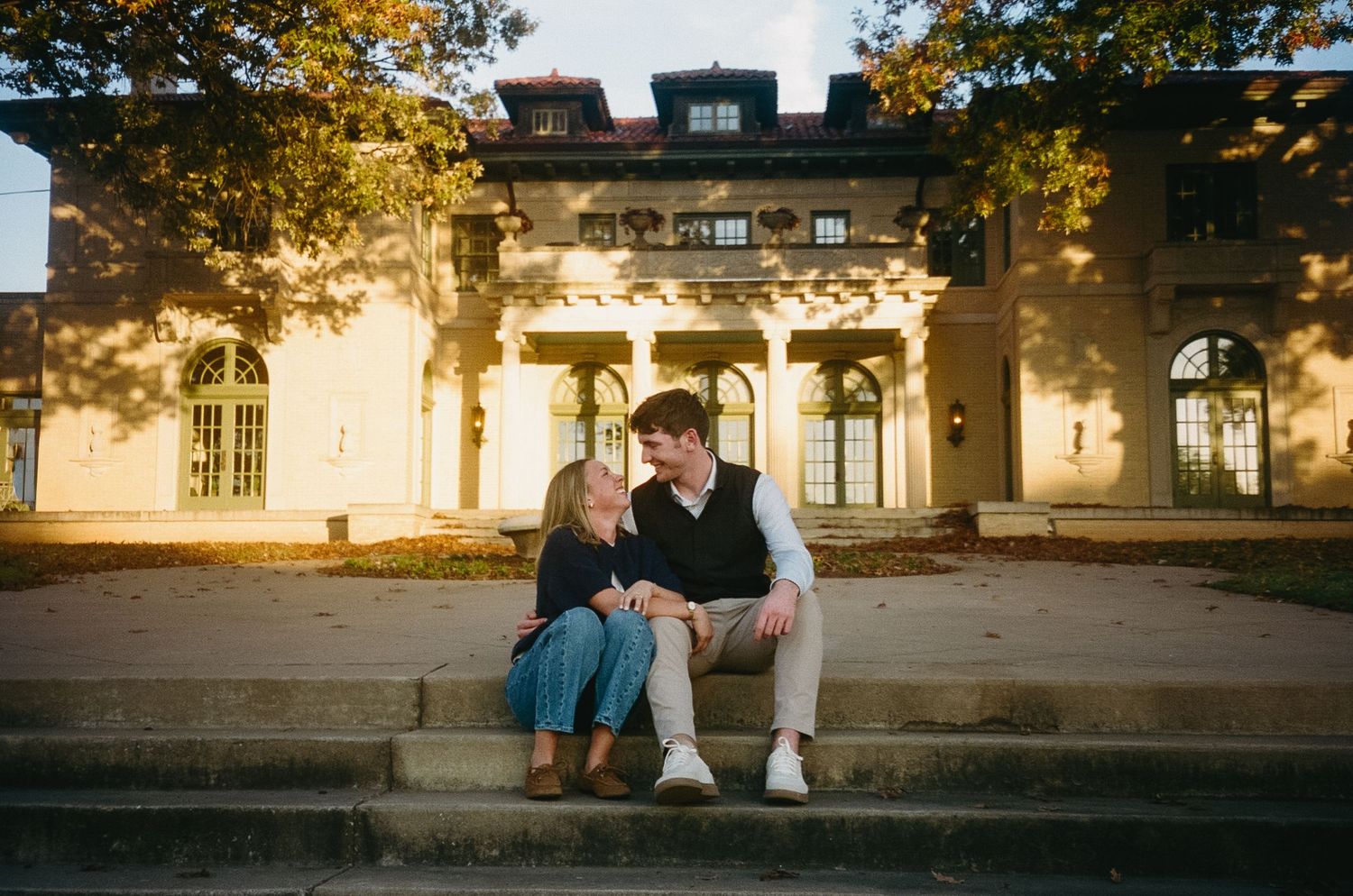 A couple sits on stone steps at sunset sharing an intimate moment in a park-like setting.
