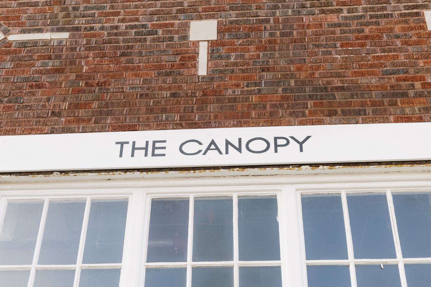 Text reading 'THE CANOPY' mounted on a red brick building above white windows.