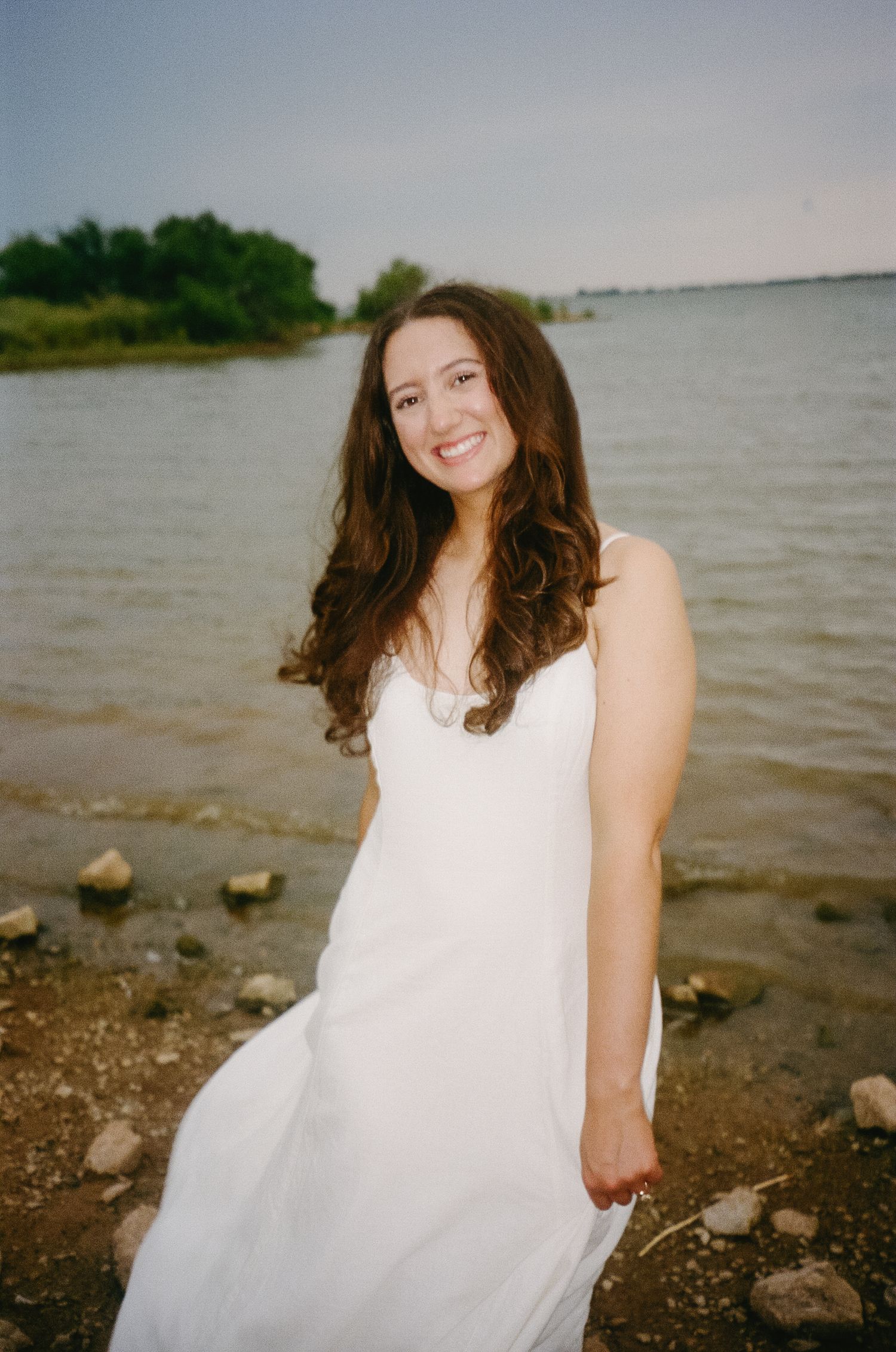 Person in a flowing white dress stands by a rocky shoreline with water and trees in the background.
