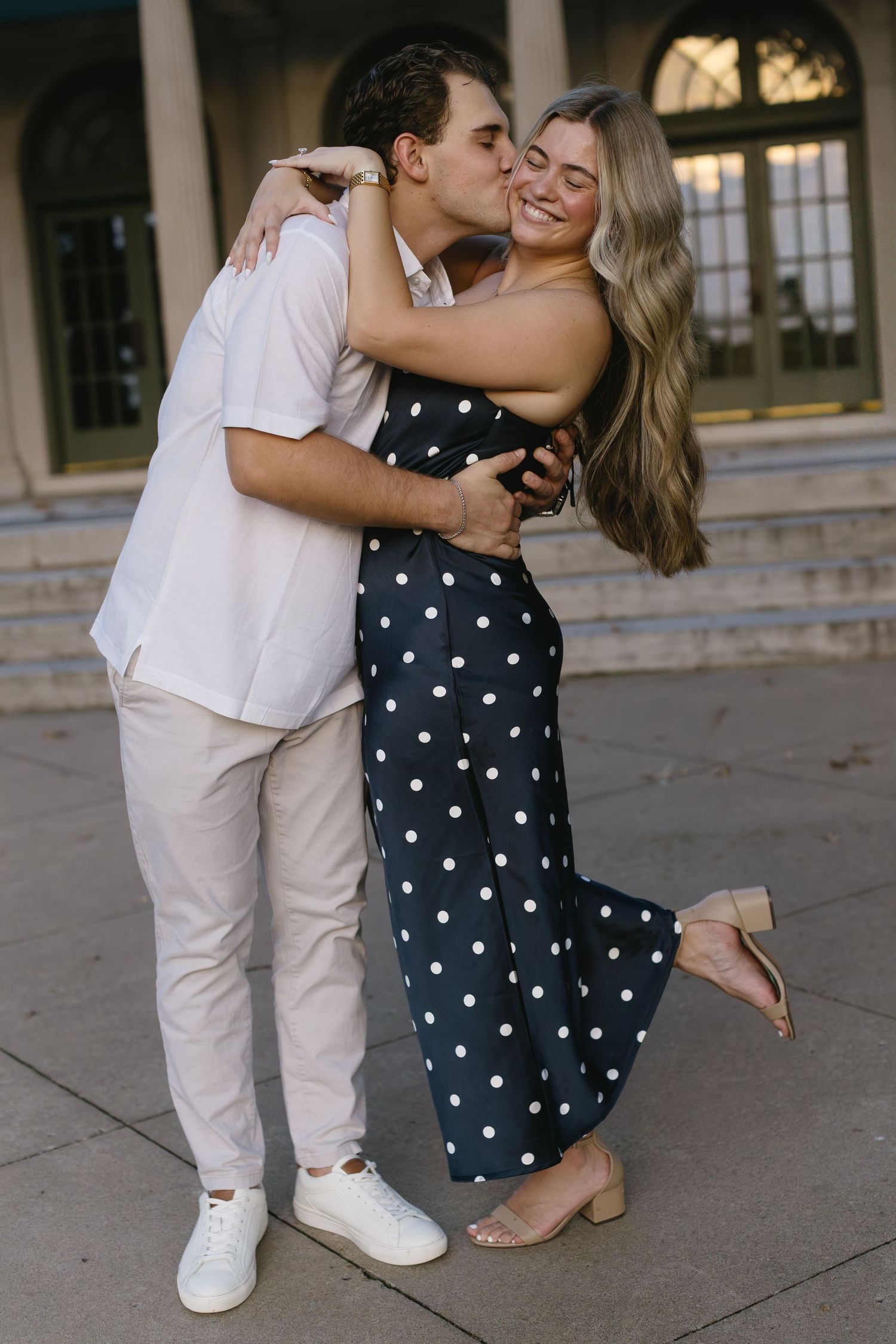 A couple embraces outdoors, with her wearing a polka dot dress and him in casual light-colored attire.
