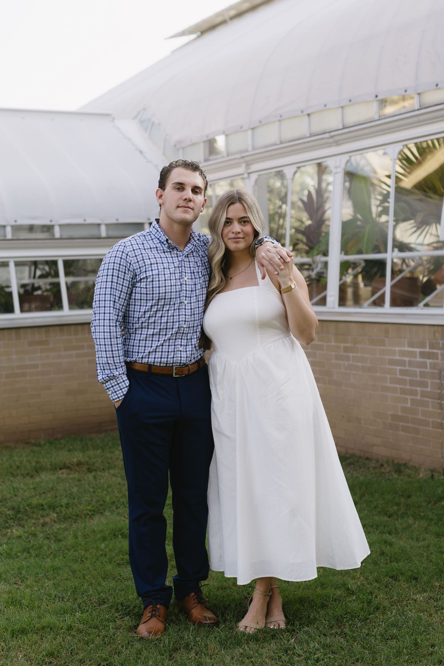 A sweet couple embracing outdoors near a glass-walled conservatory.