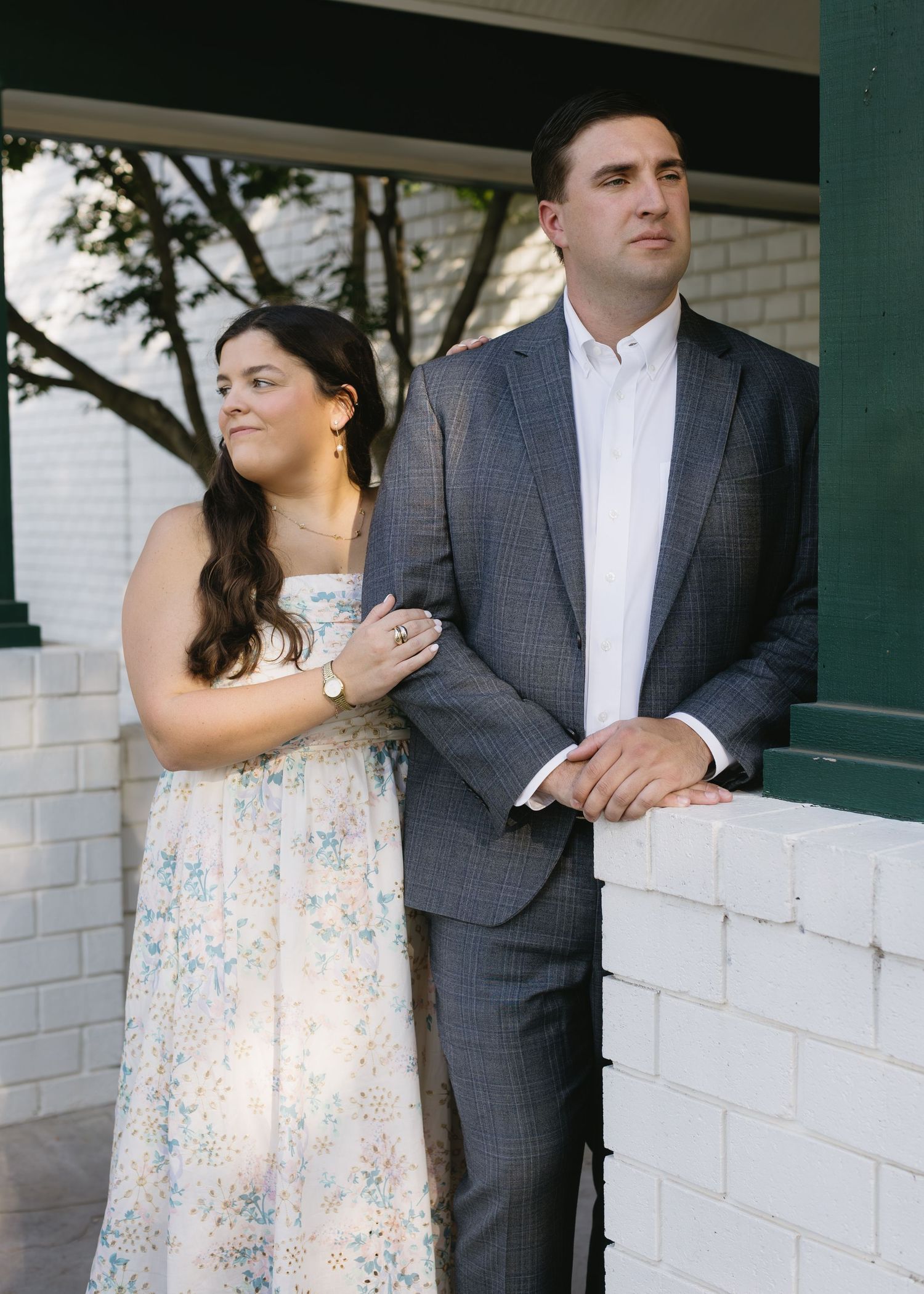 A couple stands at a white brick wall, the woman wearing a floral dress and the man in a grey suit and white shirt.