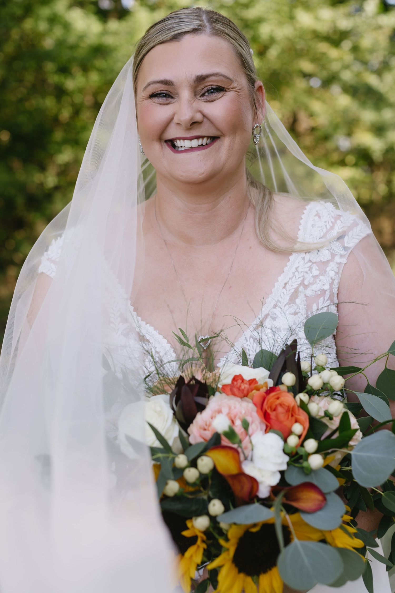 Beautiful bridal bouquet featuring sunflowers, coral roses, and eucalyptus leaves against outdoor greenery.