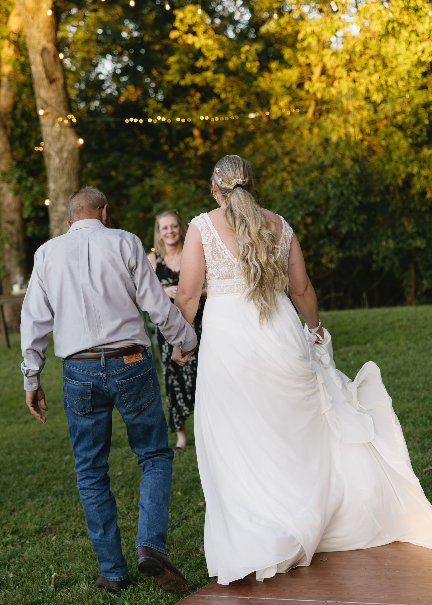 Wedding guests and a bride in white walk across a wooden deck in an outdoor garden setting.