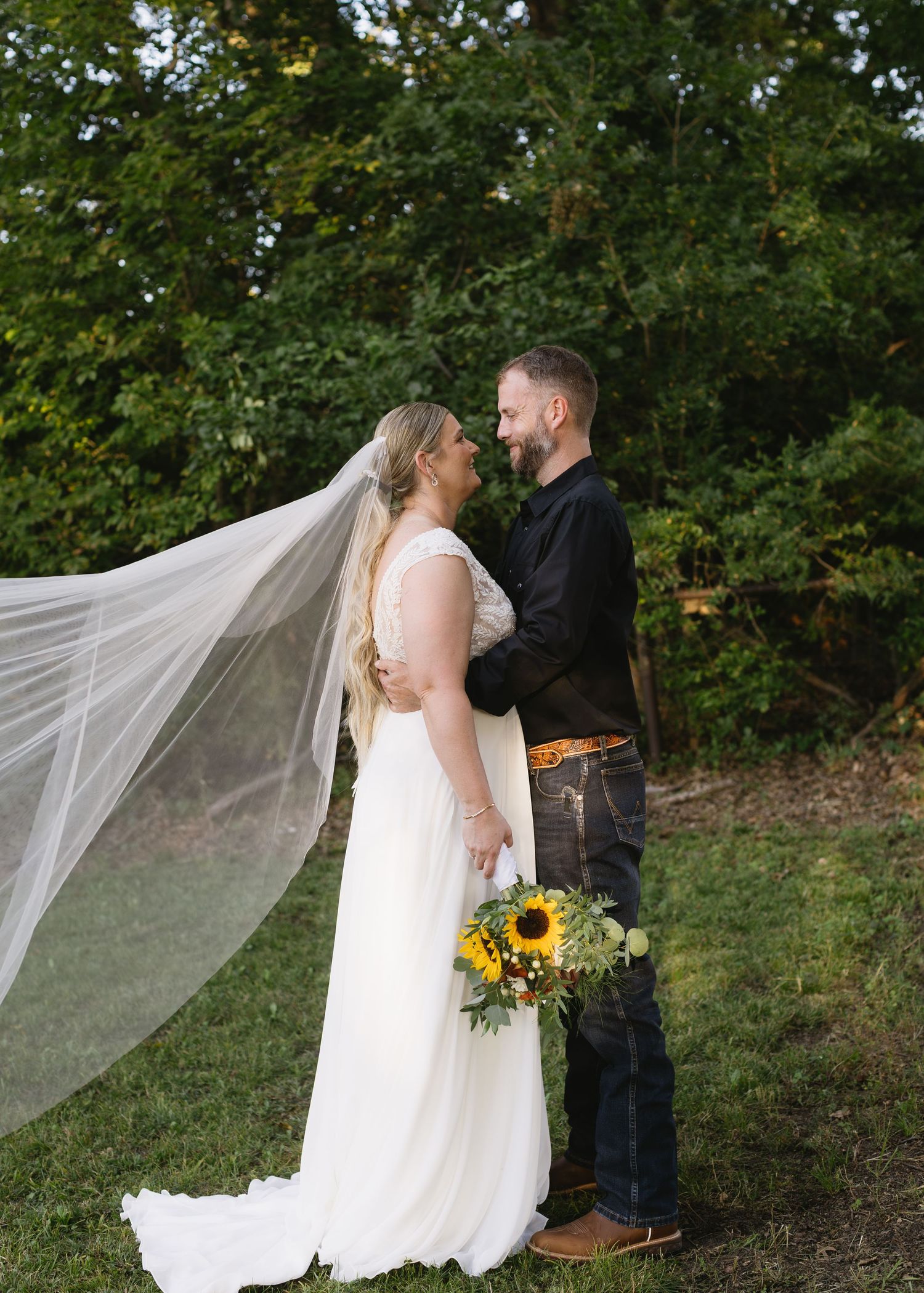 Wedding couple in outdoor setting with flowing veil and sunflower bouquet.