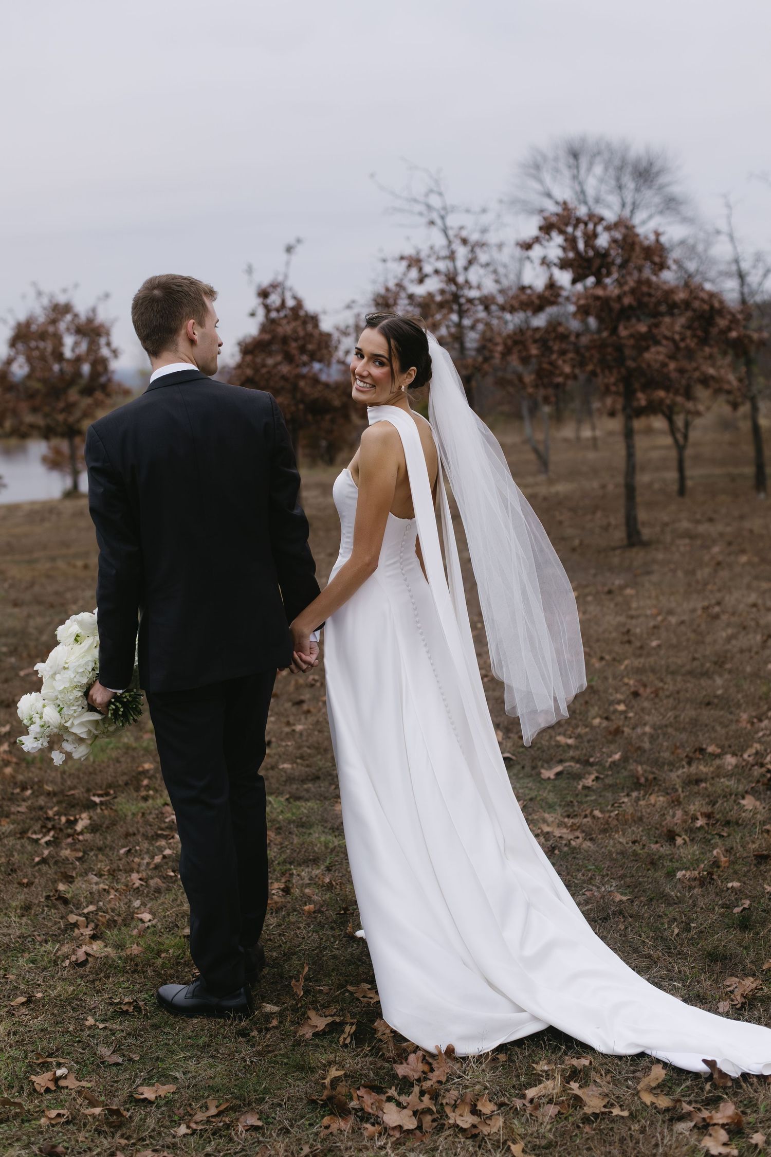 Bride in flowing white dress and groom in black suit walk through autumn field with long veil trailing behind.