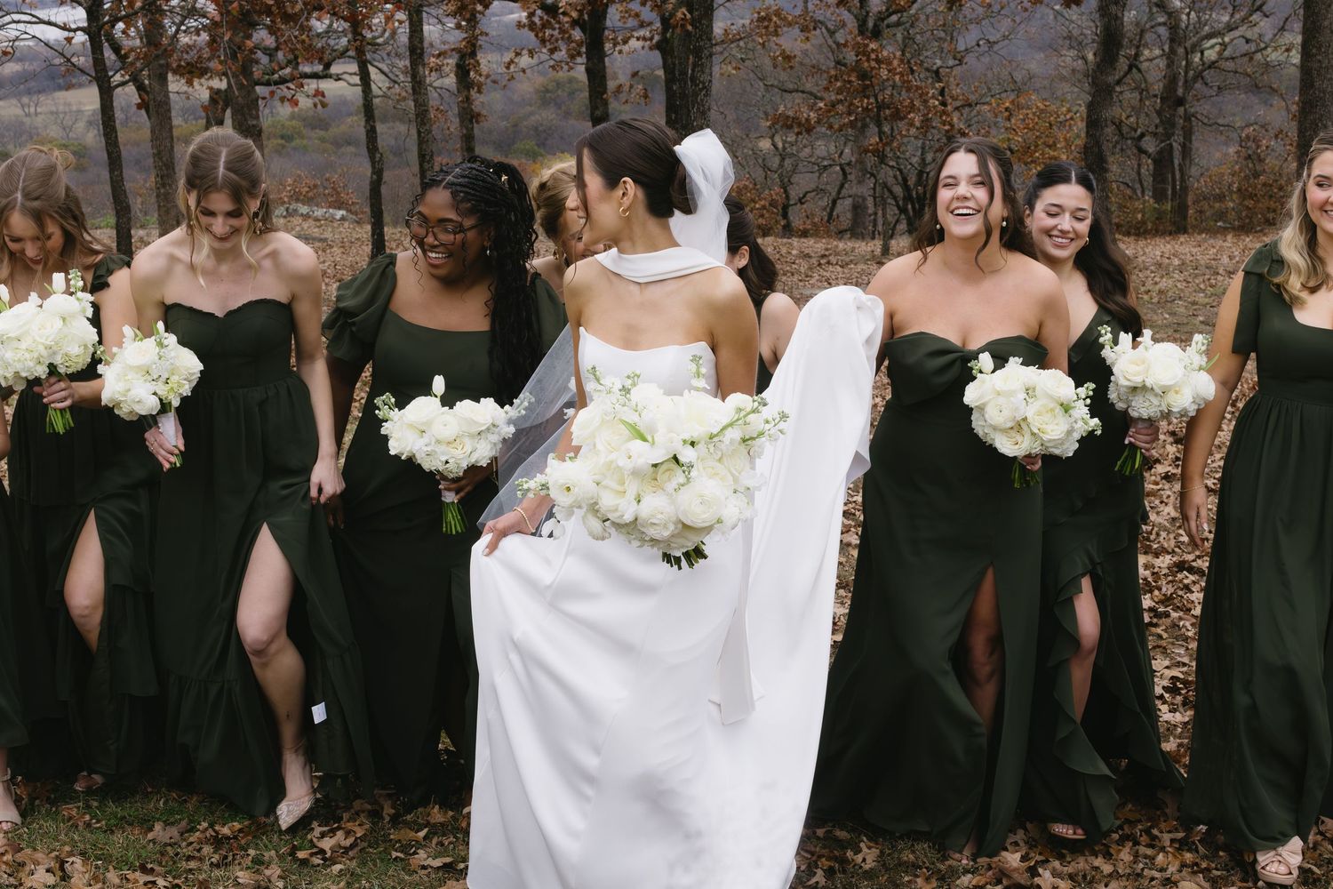 A bridal party poses in dark gowns holding white bouquets against fall foliage.
