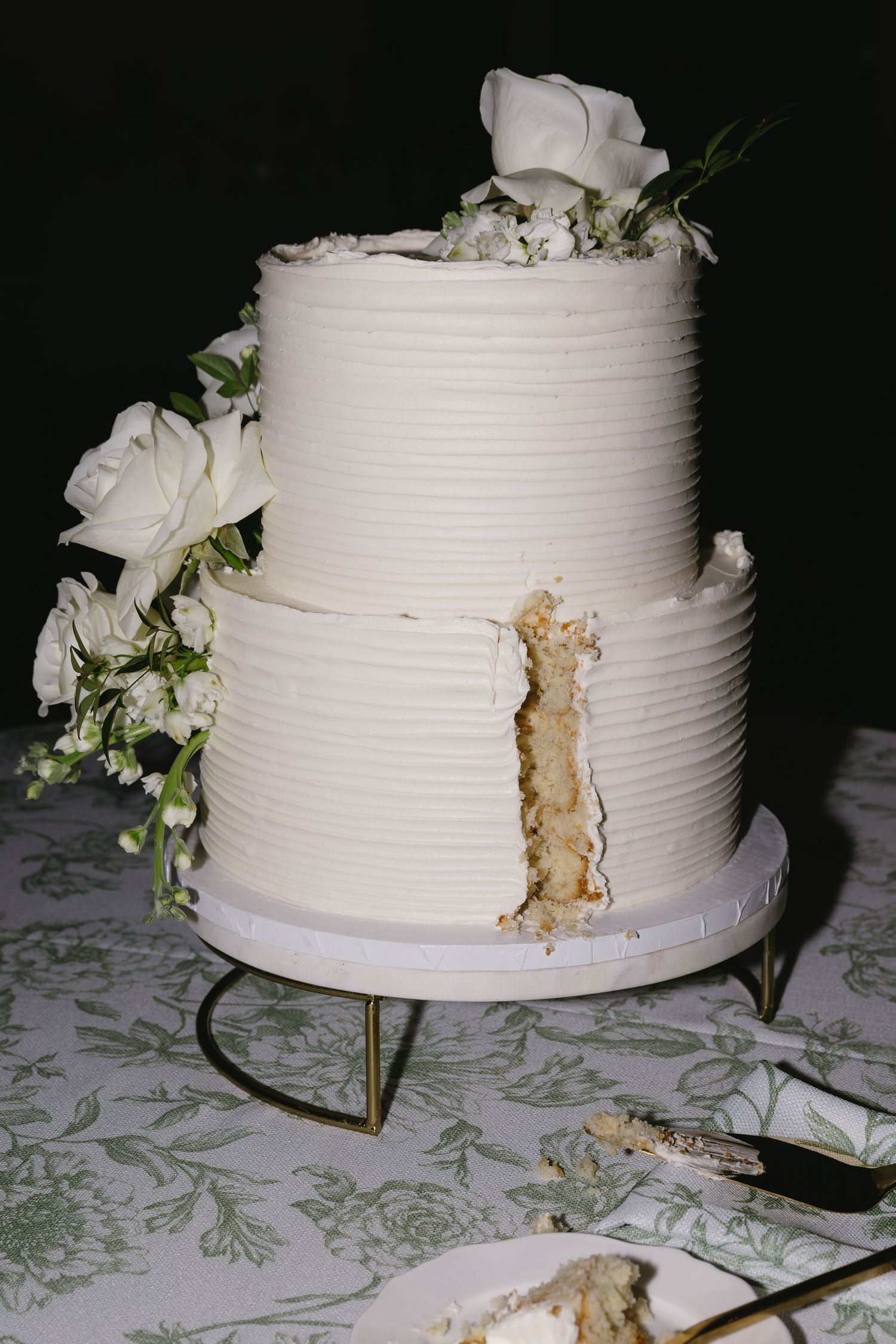 White wedding cake with textured frosting and floral decorations on a patterned tablecloth.