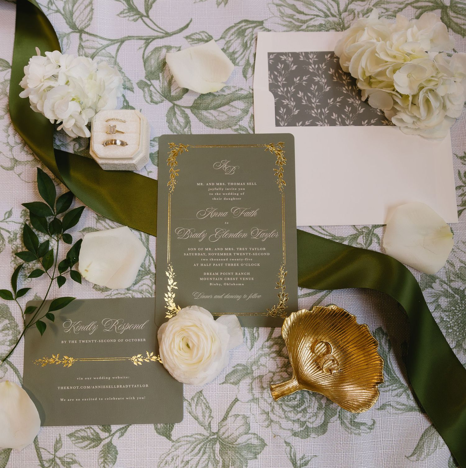 Elegant gray and gold wedding invitations styled with white roses, green ribbon and floral decor on a patterned surface.