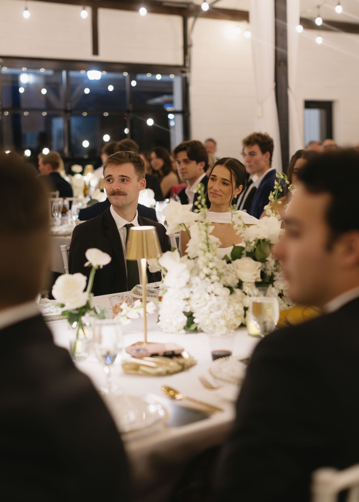 Wedding reception dinner with guests seated at elegantly decorated tables under ambient lighting.