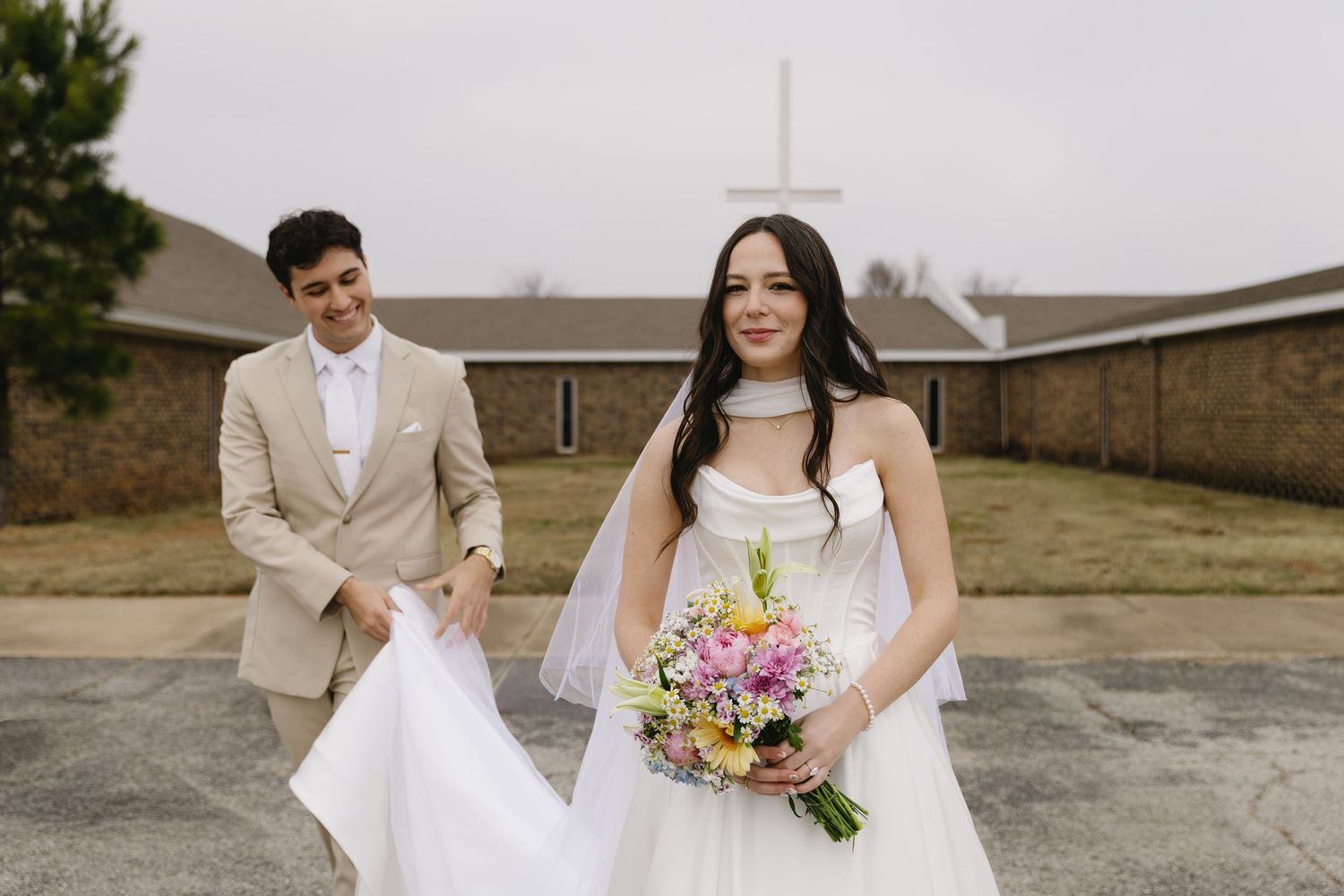 A wedding couple stands together outdoors in elegant white attire with a rustic building in the background.