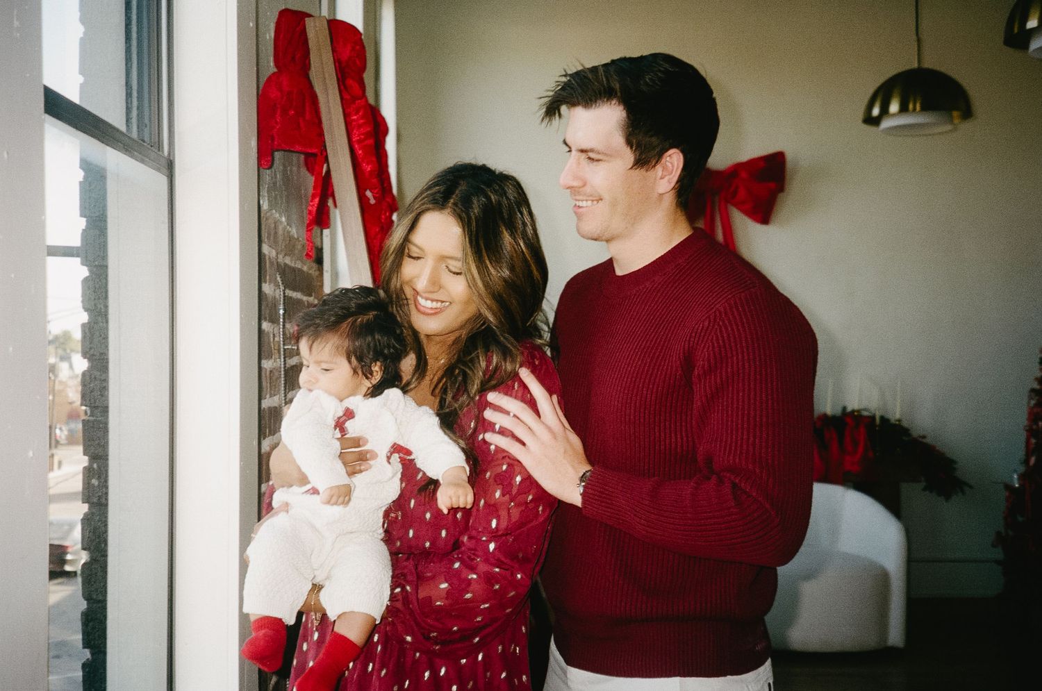 A couple in red sweaters share a tender moment near a bright window while holding a small baby.