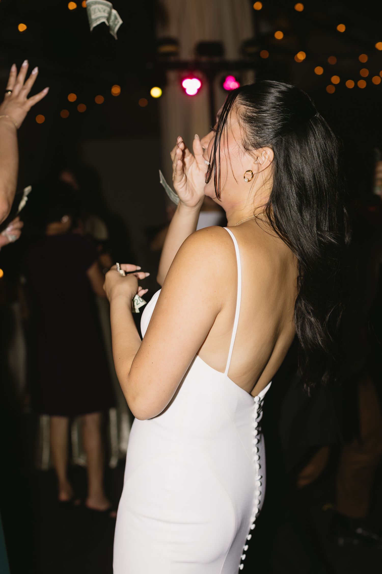 A guest in a white dress dances joyfully at a wedding reception with twinkling lights in background.