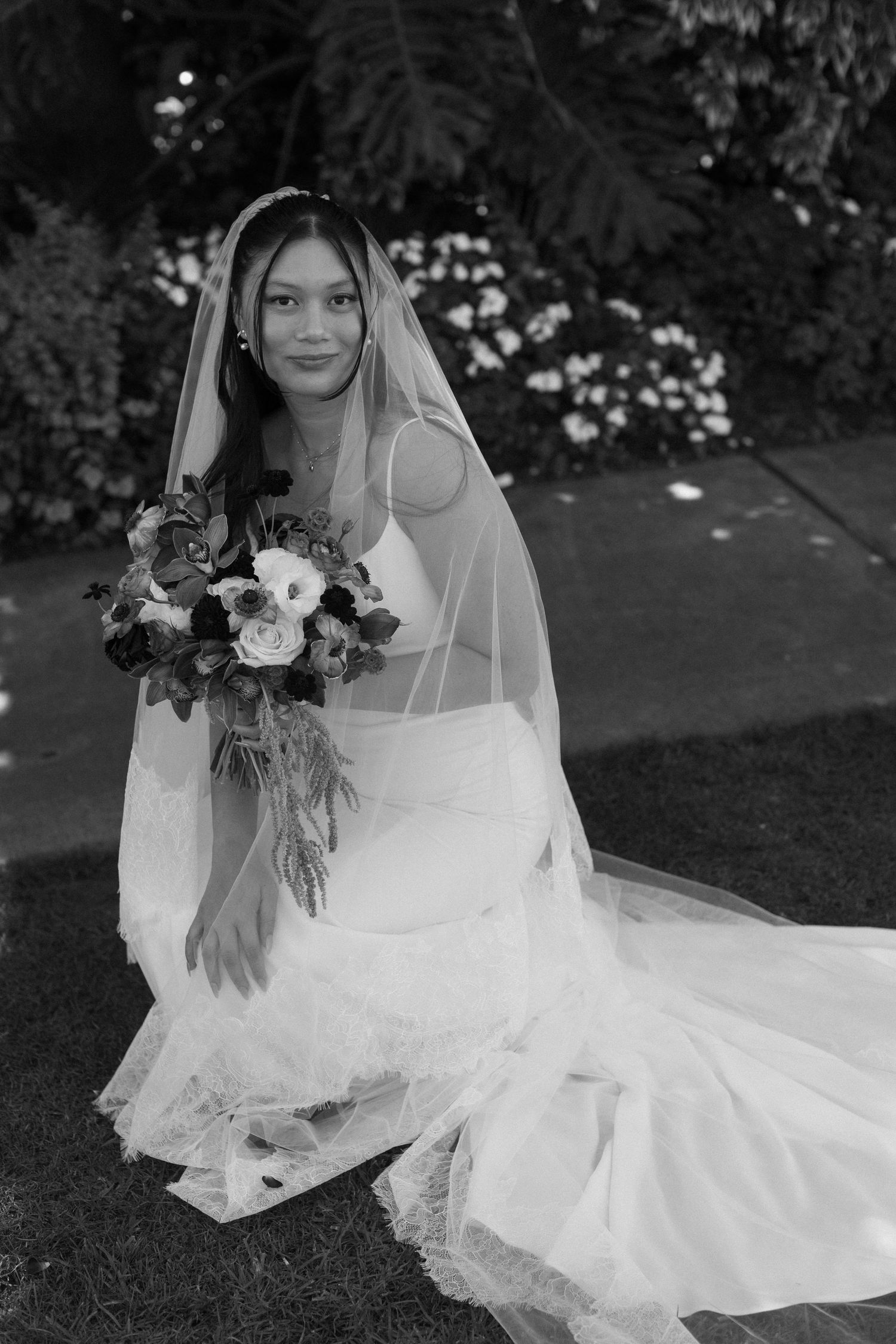 A bride in a white wedding dress poses gracefully on the ground with a bouquet of dark and light flowers.