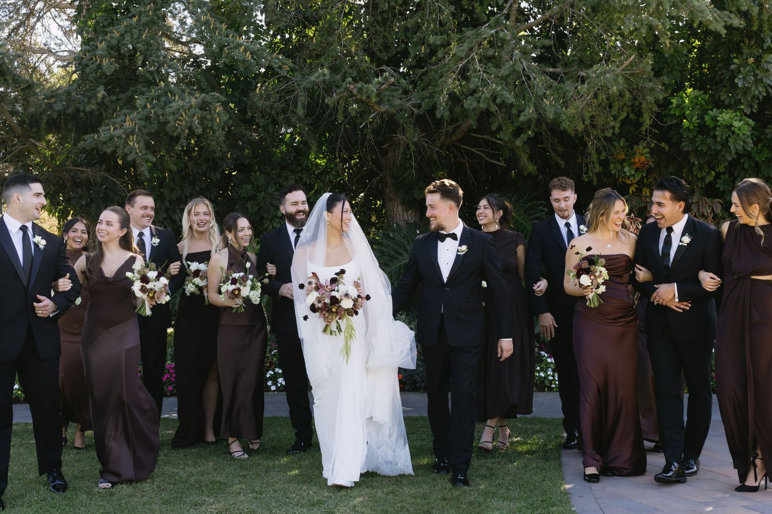 A wedding party poses outdoors with bridesmaids in brown dresses and groomsmen in black tuxedos.