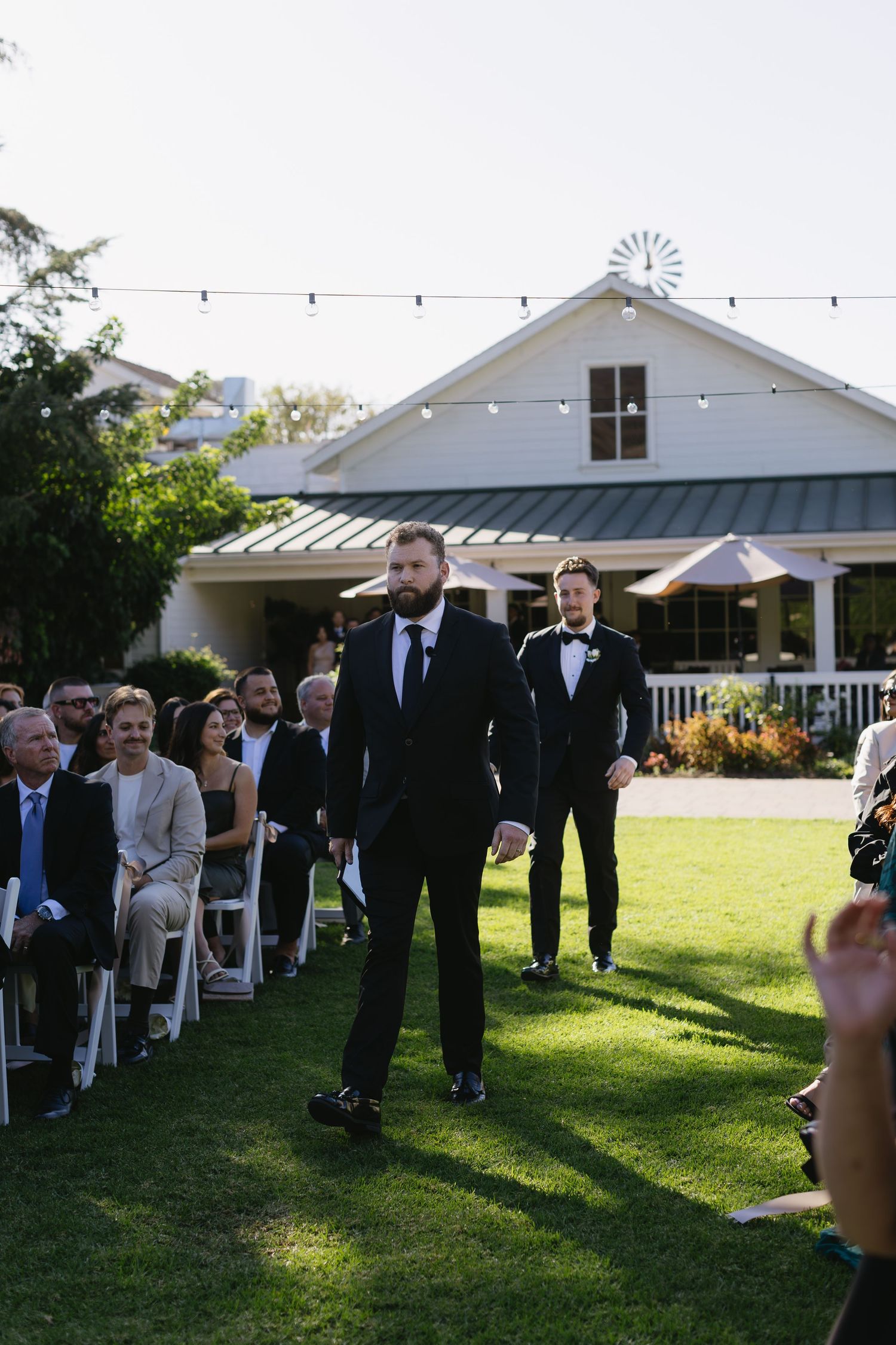Wedding guests gathering on a lawn outside a white farmhouse venue with string lights overhead.