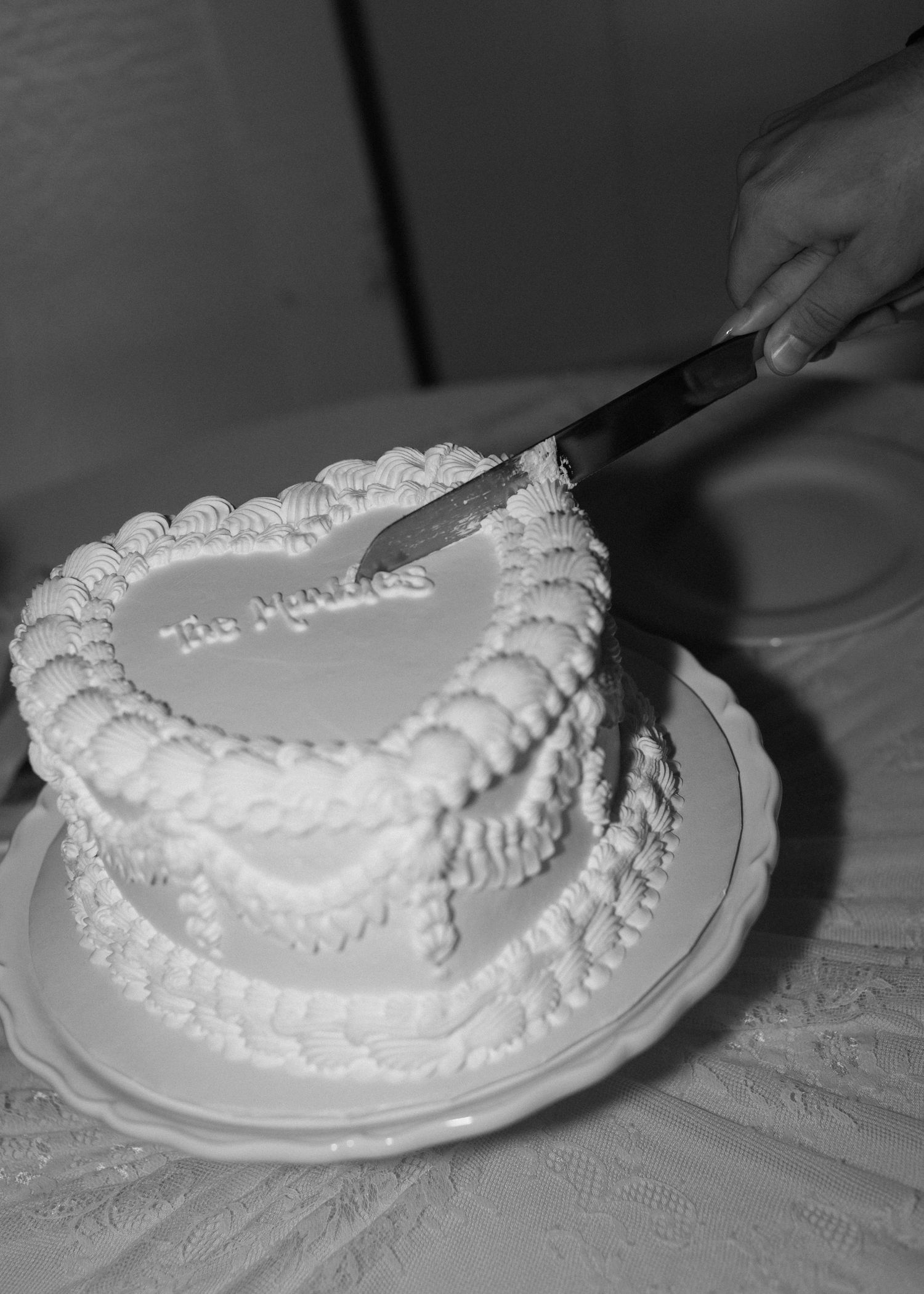 A heart-shaped white wedding cake being cut with a silver knife against a lace tablecloth.