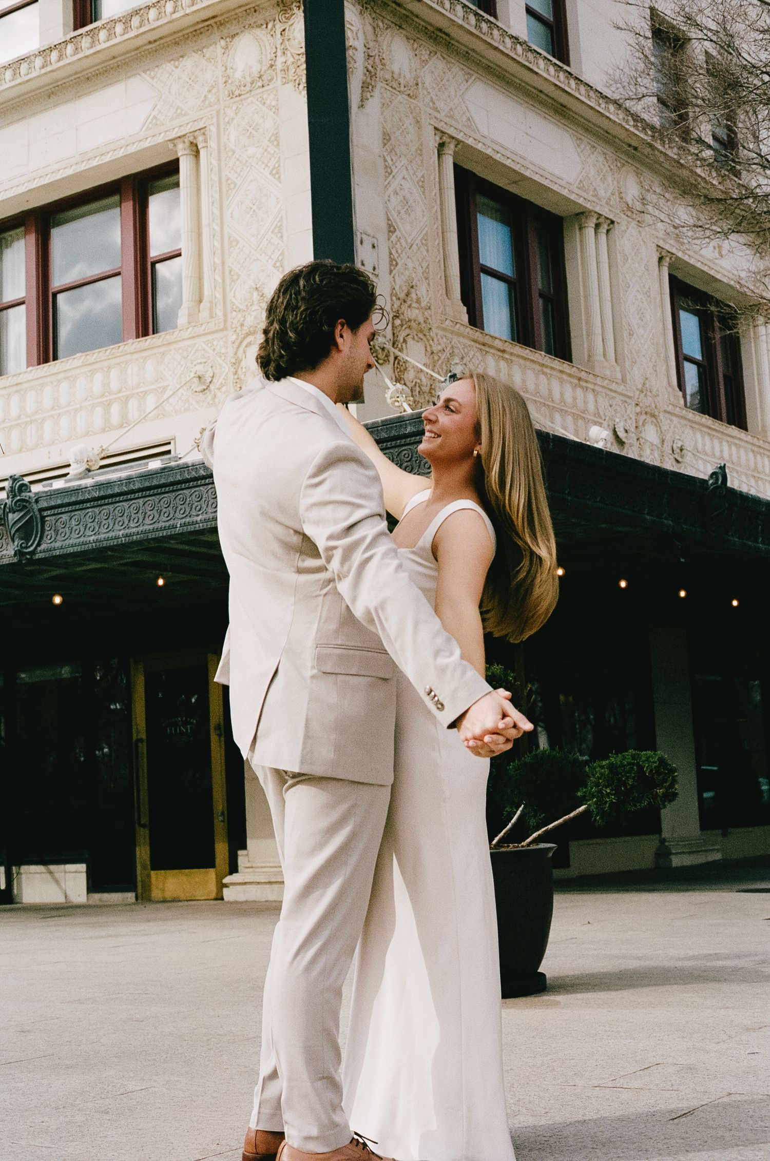 A couple in formal white attire dances together outside a historic building.