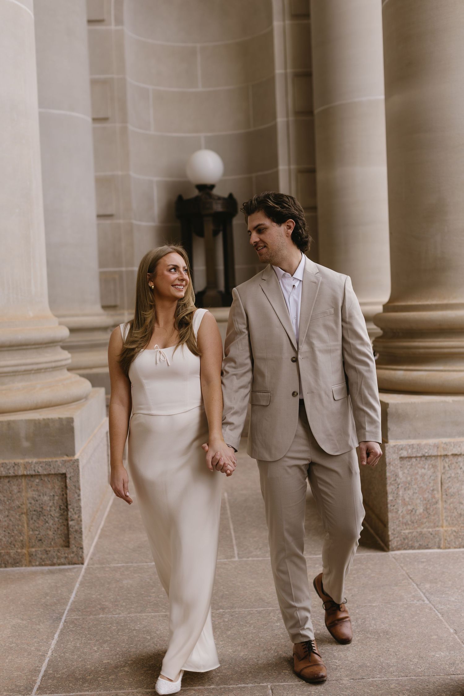 A couple in formal attire walks together between elegant stone columns, wearing beige and white.