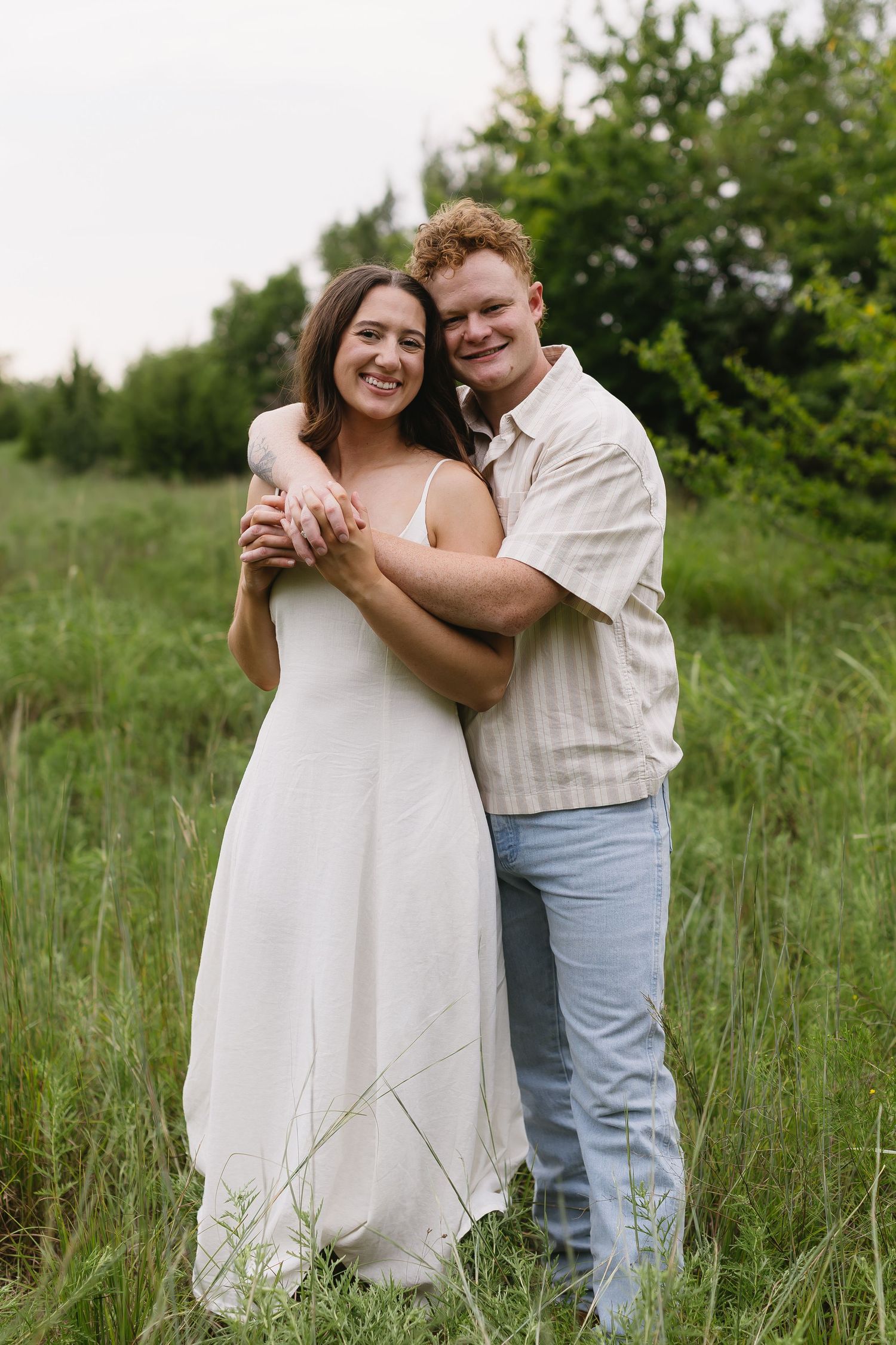A couple embraces joyfully in a sunlit meadow with green foliage in the background.