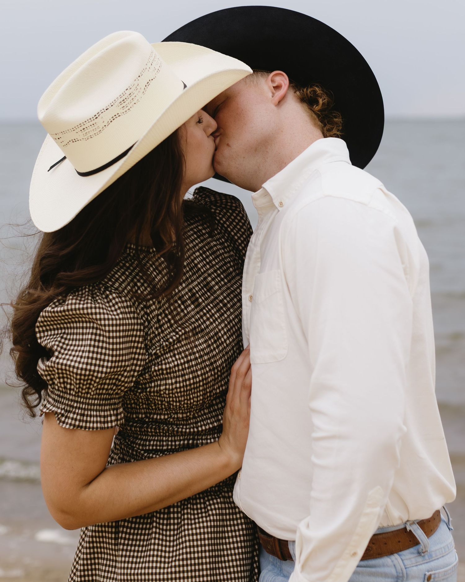A romantic couple in western attire share intimate moments by the water's edge during a photoshoot.
