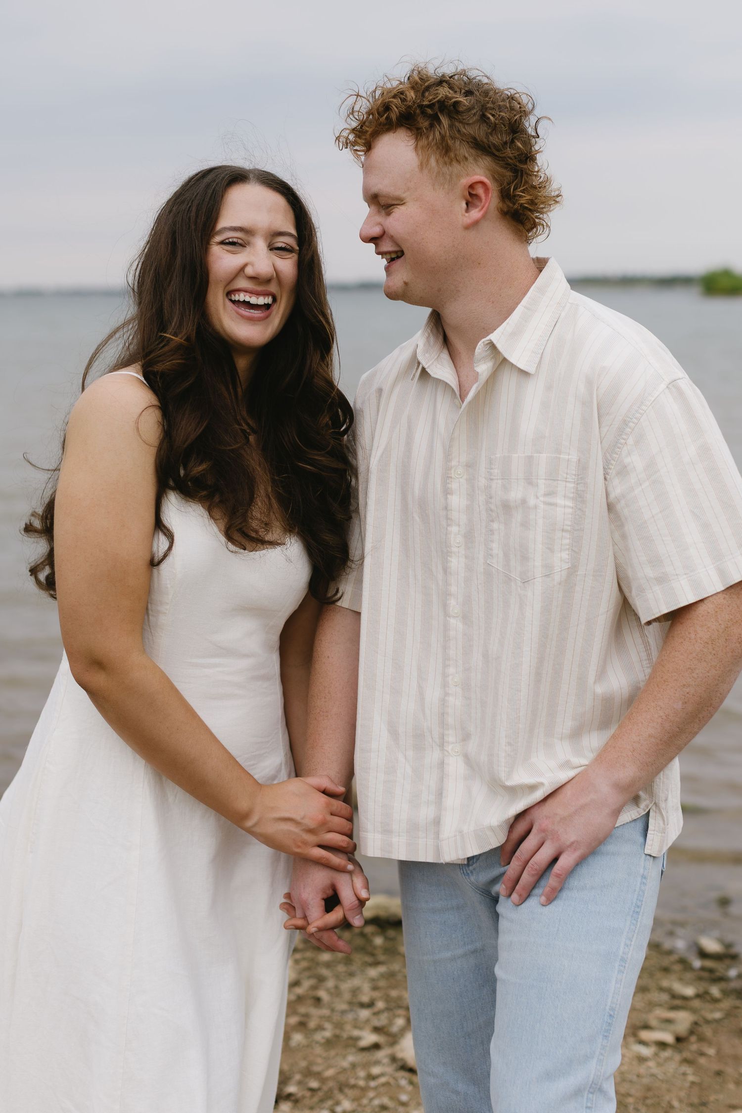 A couple laughs together during a waterfront photoshoot wearing white and denim outfits.