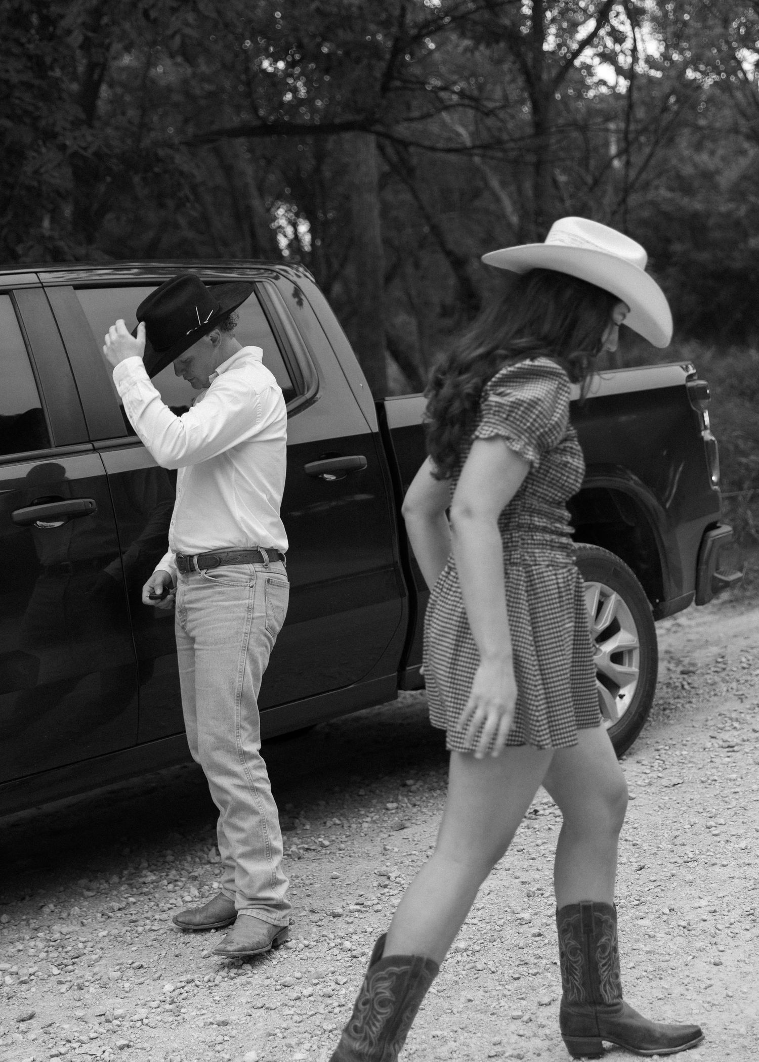 Black and white photo of two people in boots standing near parked cars on a gravel path.