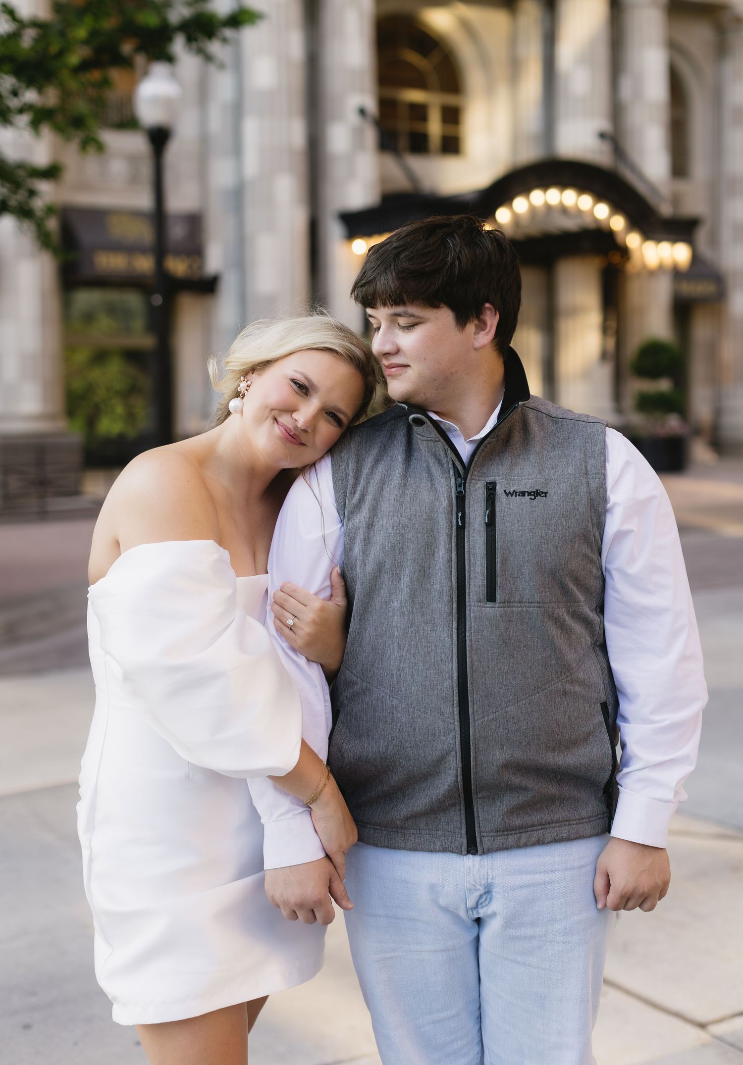 A couple in casual attire shares a tender moment while standing outdoors in an urban setting.
