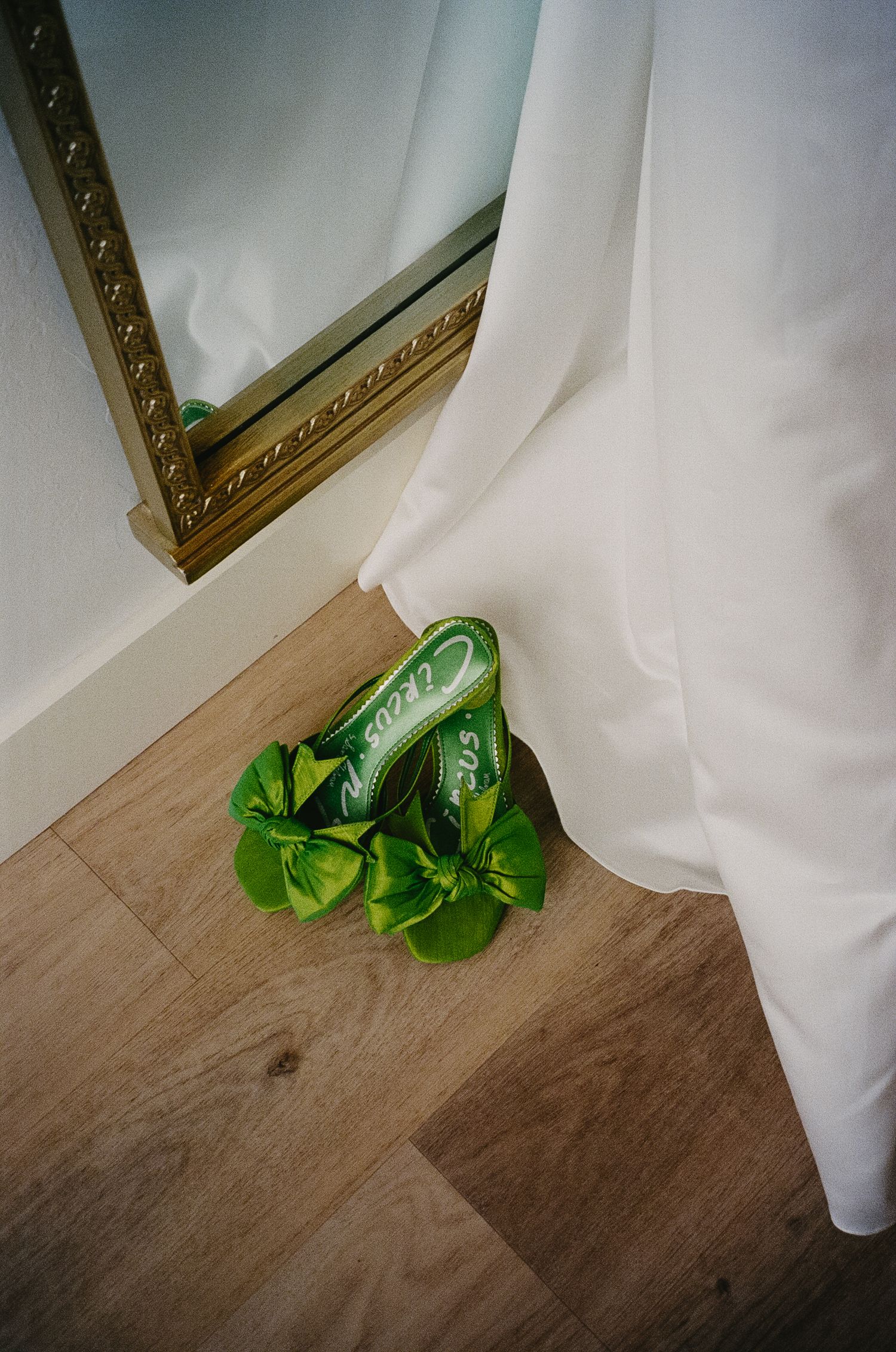 Green satin wedding shoes peek out beneath flowing white fabric on wooden flooring.