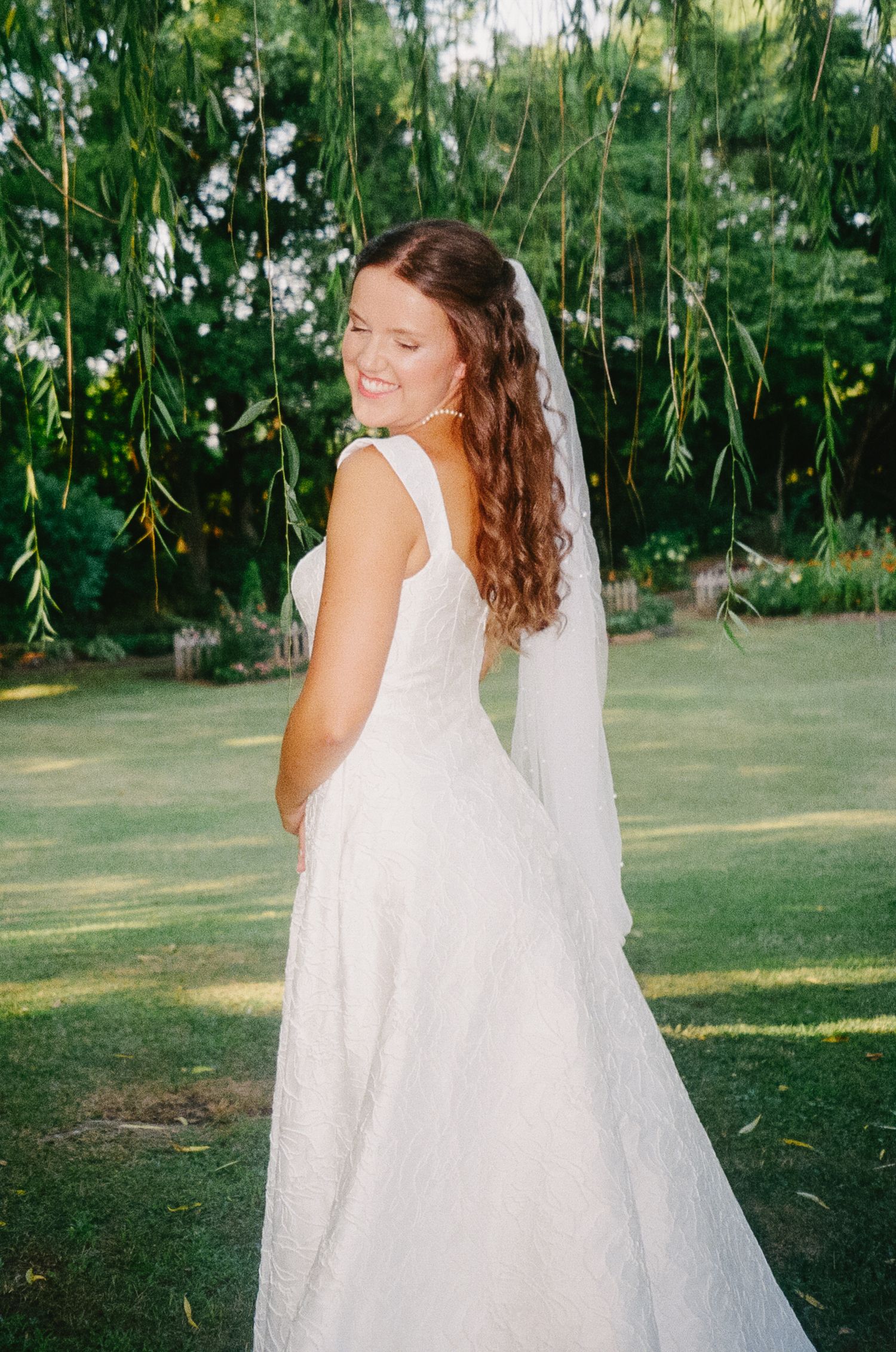 Elegant wedding dress captured in motion against lush garden greenery and hanging willow branches.
