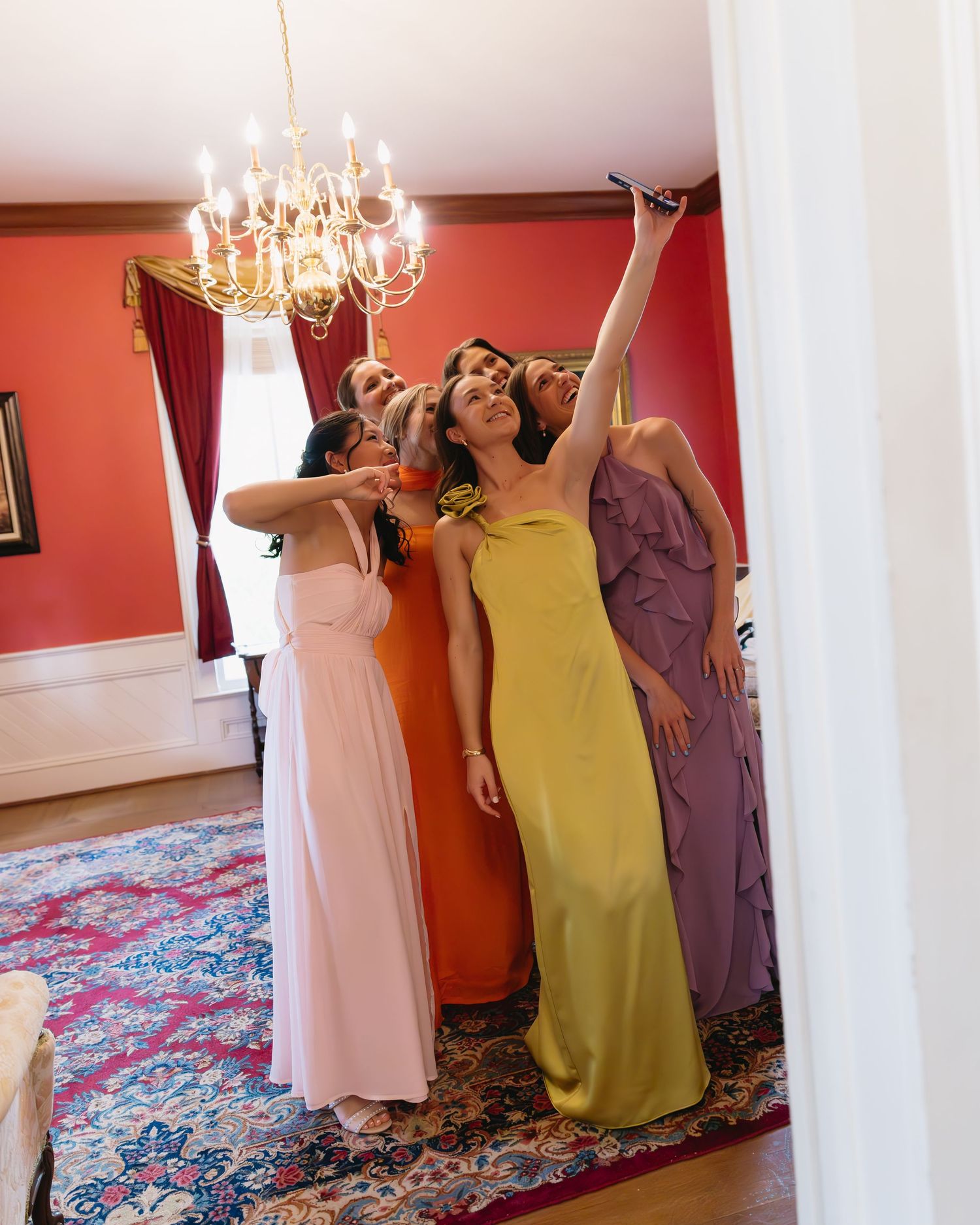 Three women in formal dresses strike playful poses under an elegant crystal chandelier in a room with coral-colored walls.