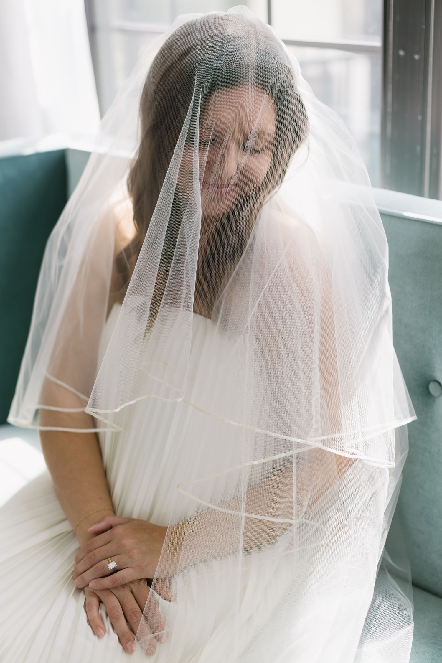 A bride in a white dress and veil sits on a teal couch looking down.