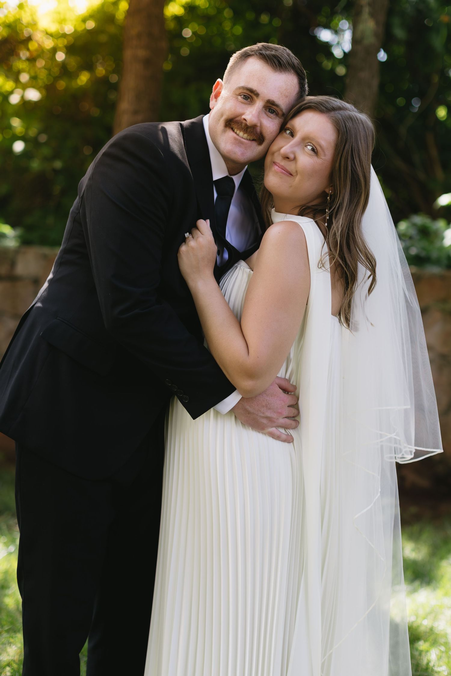 Newlywed couple embracing and laughing together in a sun-dappled garden setting.