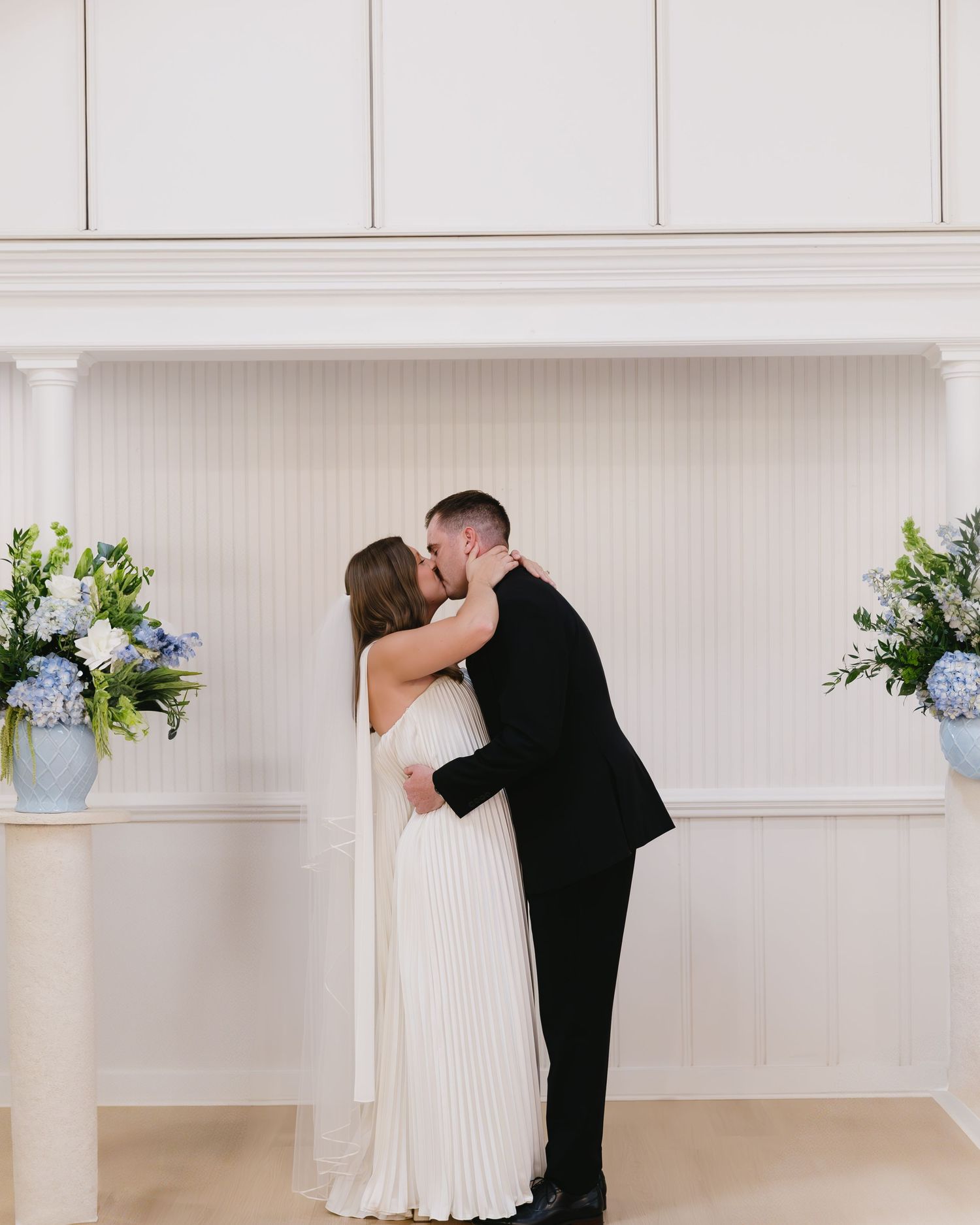 Elegant wedding couple sharing kiss in white room with floral arrangements.