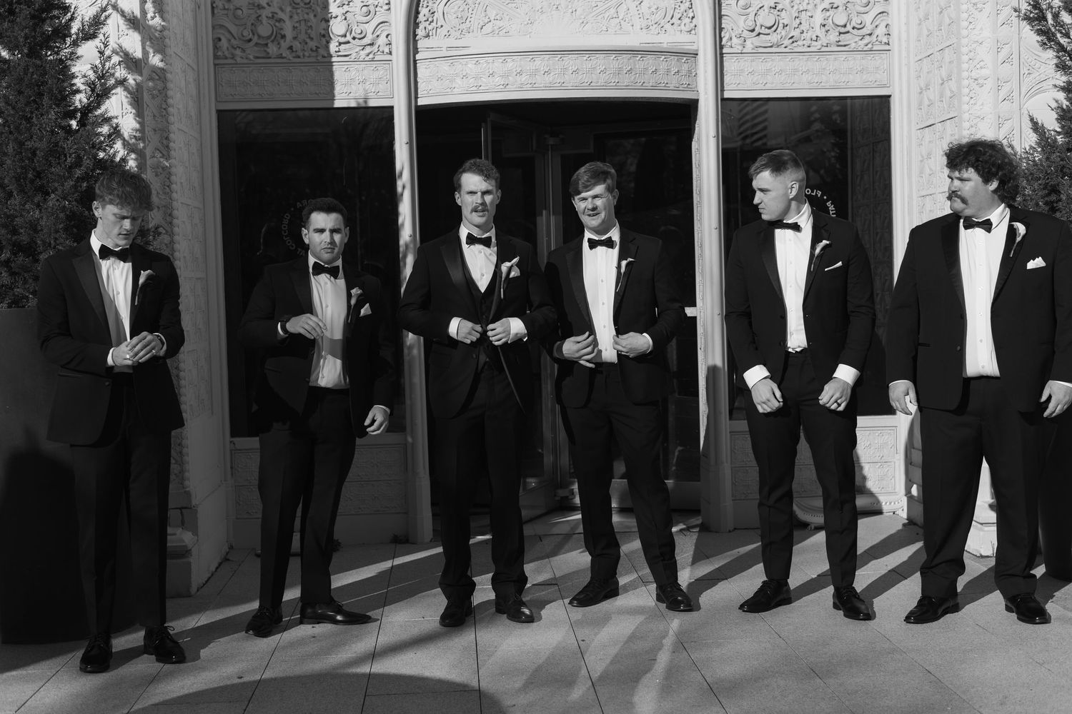 Groomsmen lined up against wall wearing classic black tuxedos in formal wedding party photo.