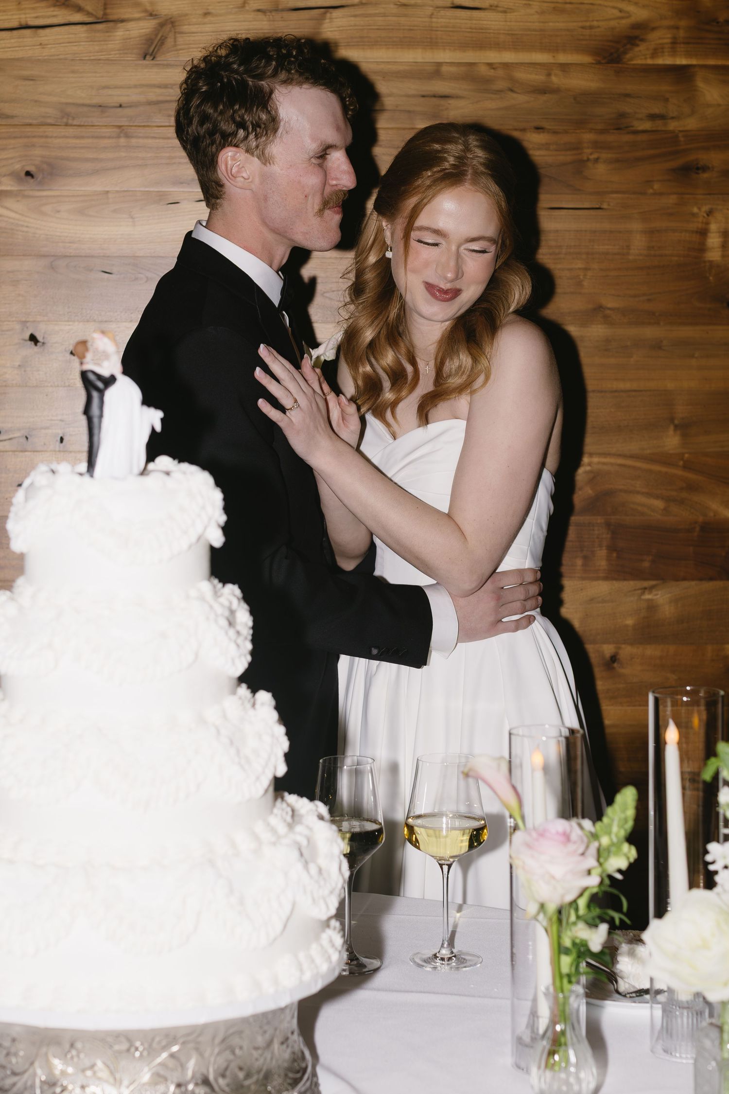 Wedding reception scene with a white cake and dancing couple against rustic wooden backdrop.