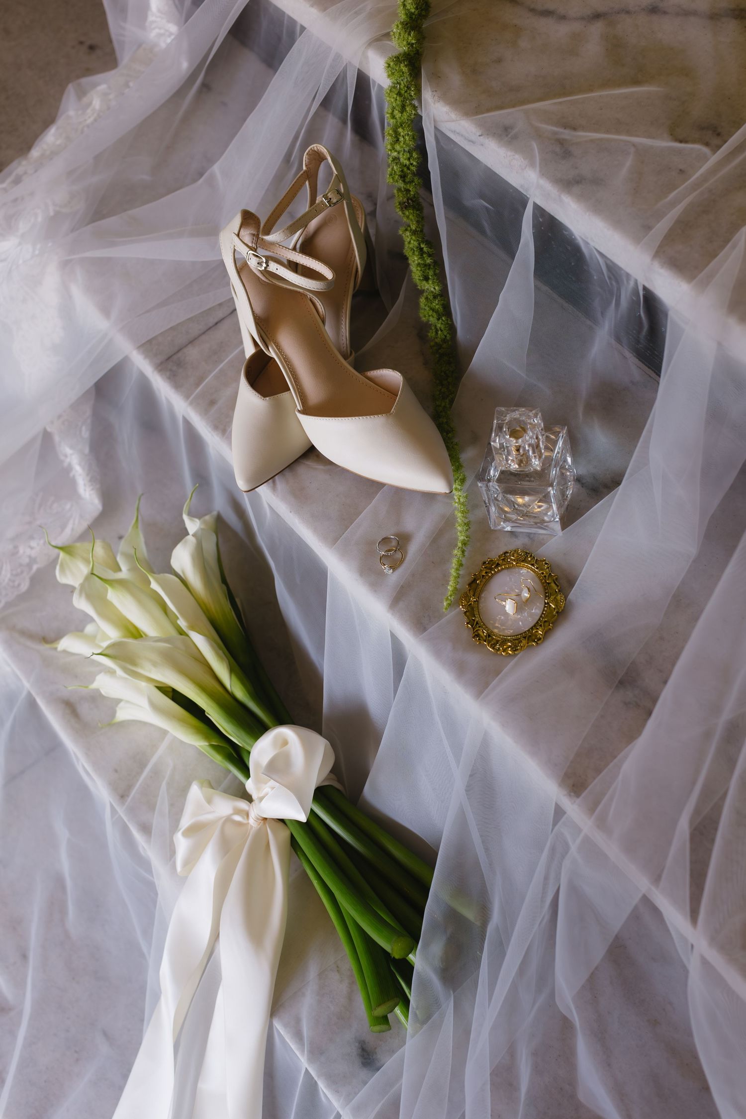 Wedding accessories including white heels, jewelry, and calla lily bouquet arranged on flowing tulle fabric.