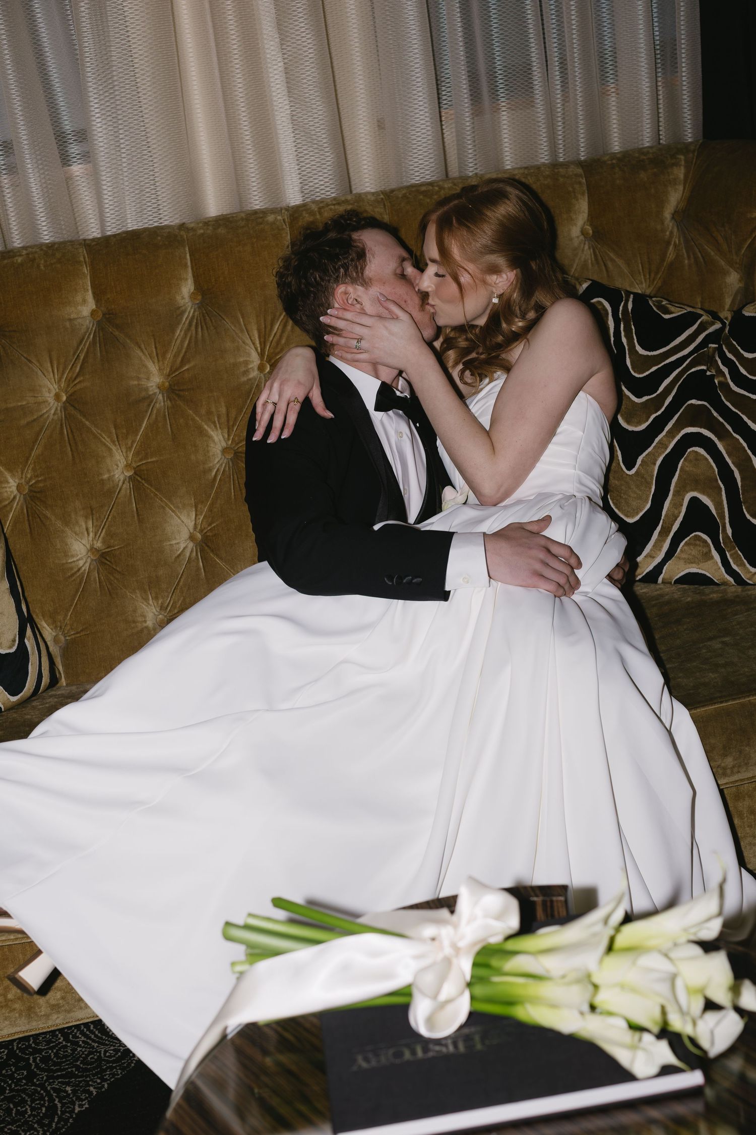 Newlyweds share intimate moment on gold velvet couch with white flowers nearby.