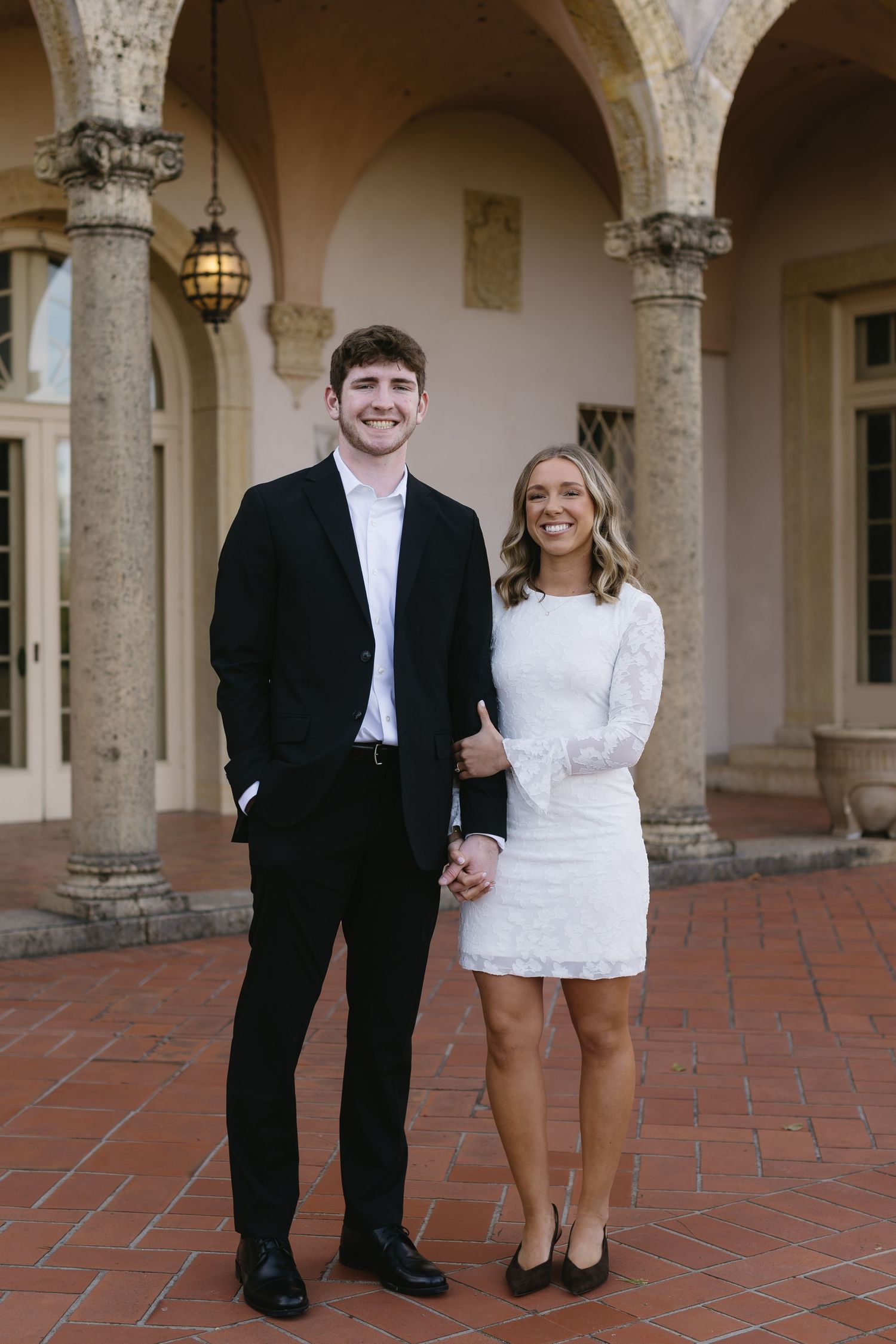 A couple in formal attire poses together in an elegant courtyard with stone columns.