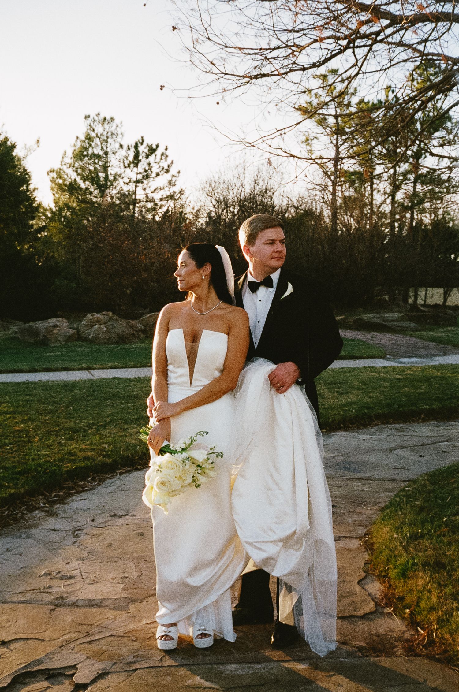 A couple in wedding attire sits on stone steps during golden hour surrounded by trees.