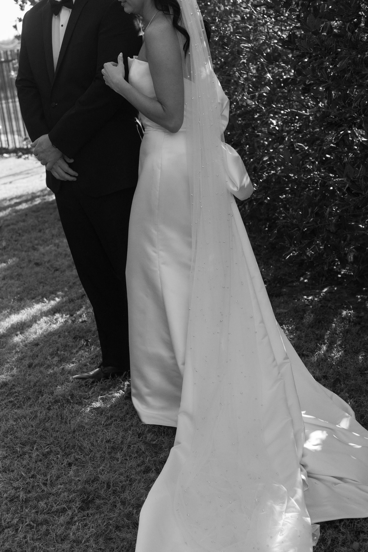 Black and white wedding photo showing elegant bridal gown with flowing train against garden backdrop.