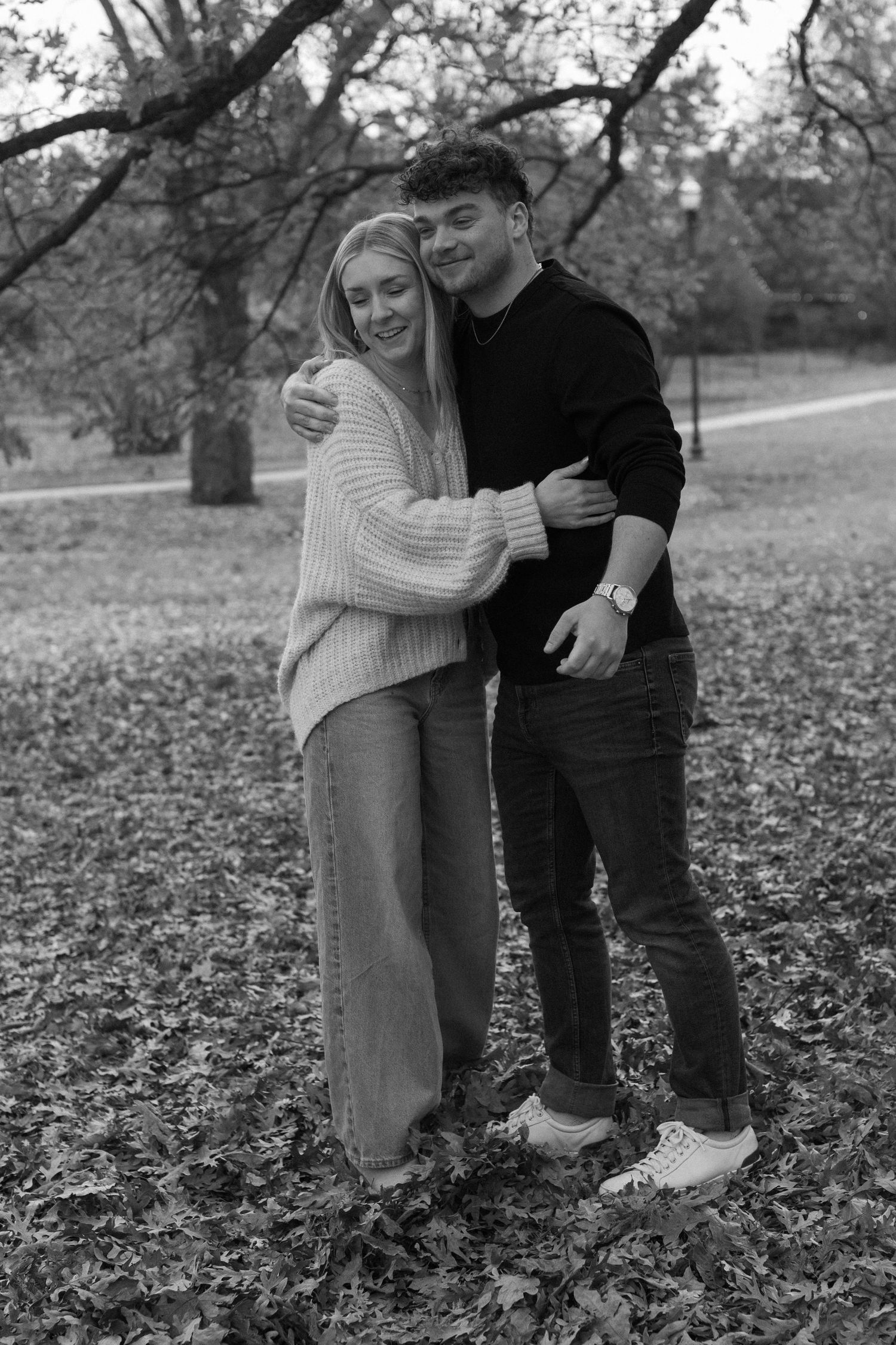 Black and white photo of a couple posing together in a leaf-covered autumn landscape.