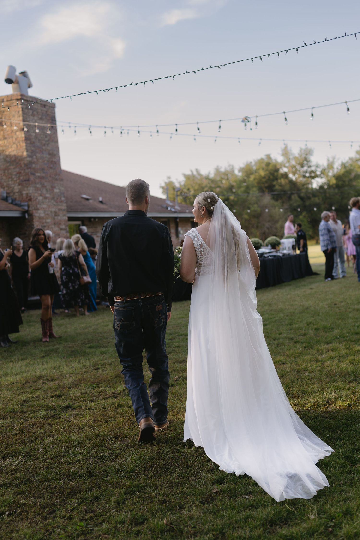 Wedding reception under string lights with guests mingling on lawn at dusk.