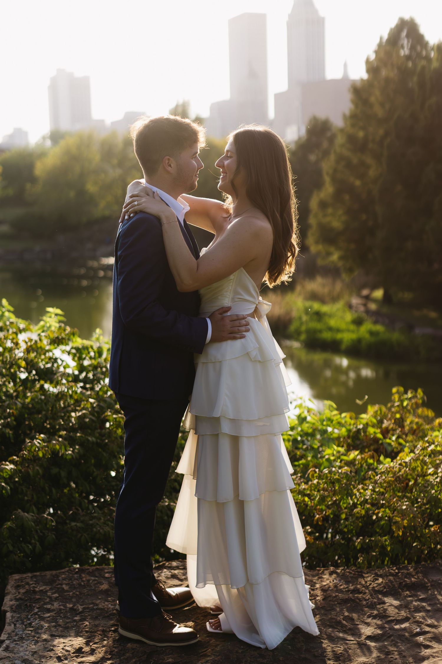 A couple embraces during golden hour with cityscape and garden scenery in the background.